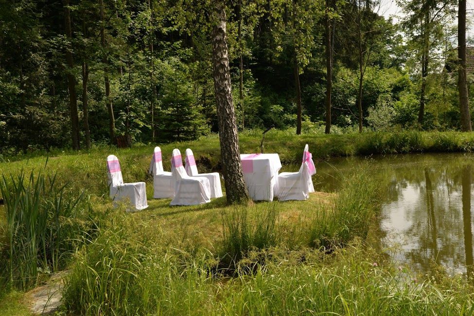 Stühle und ein Tisch mit weißen Hussen auf einer grünen Wiese neben einem Teich, umgeben von Wald.