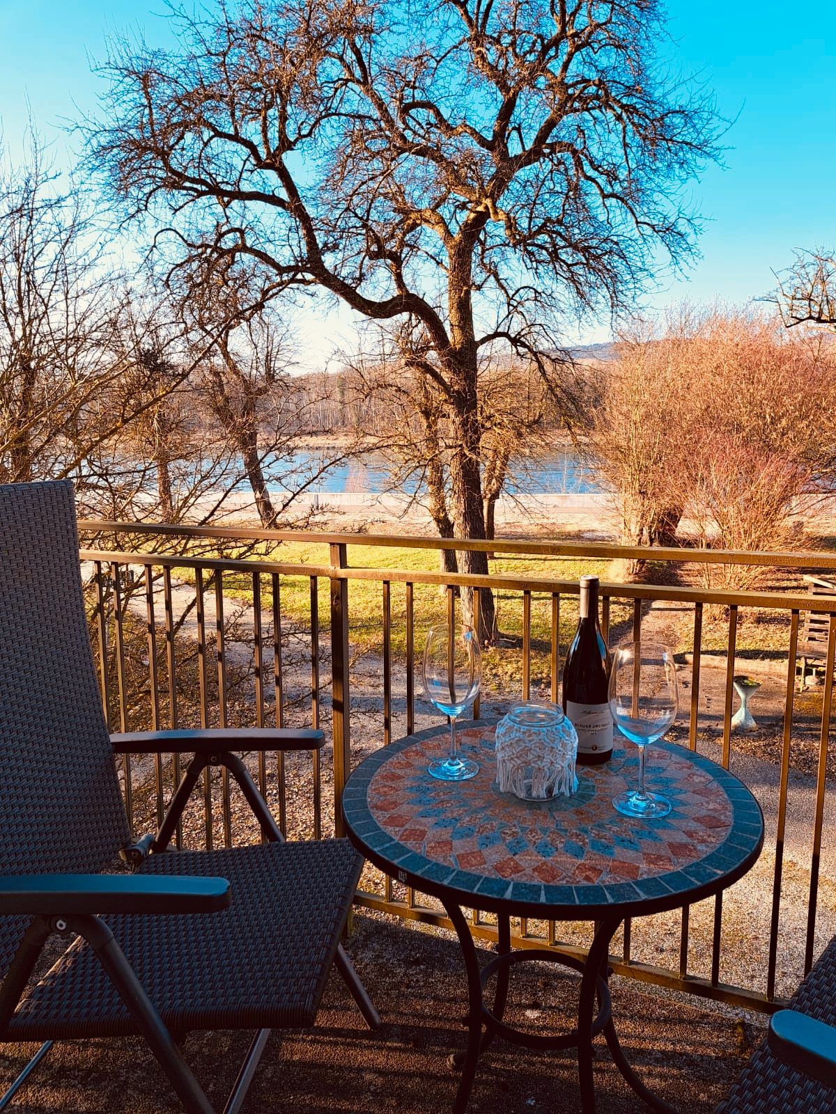 Balkon mit Tisch, zwei Weingläsern und Weinflasche, Blick auf Bäume und Fluss.