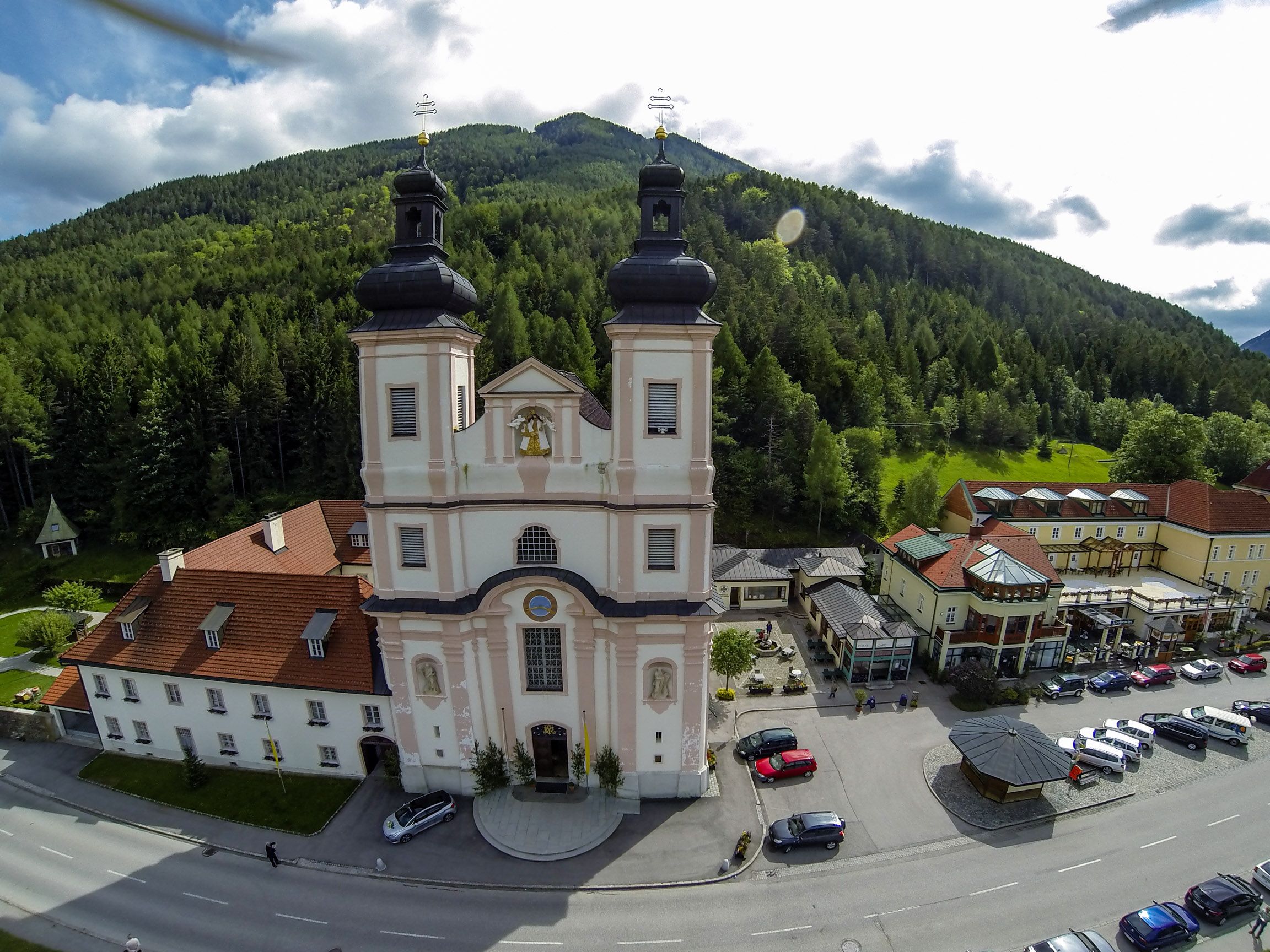 Luftaufnahme einer Kirche mit zwei Türmen, umgeben von Gebäuden und Wald.