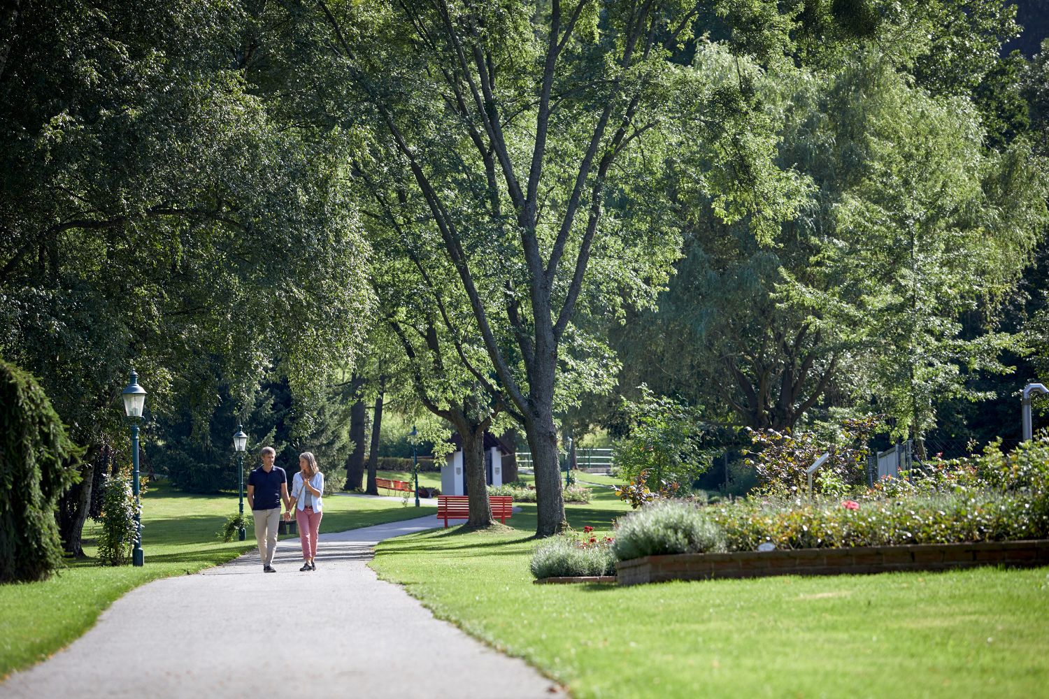 Ein Paar spaziert auf einem Weg im Kurpark Bad Schönau, umgeben von grünen Bäumen und gepflegten Blumenbeeten.