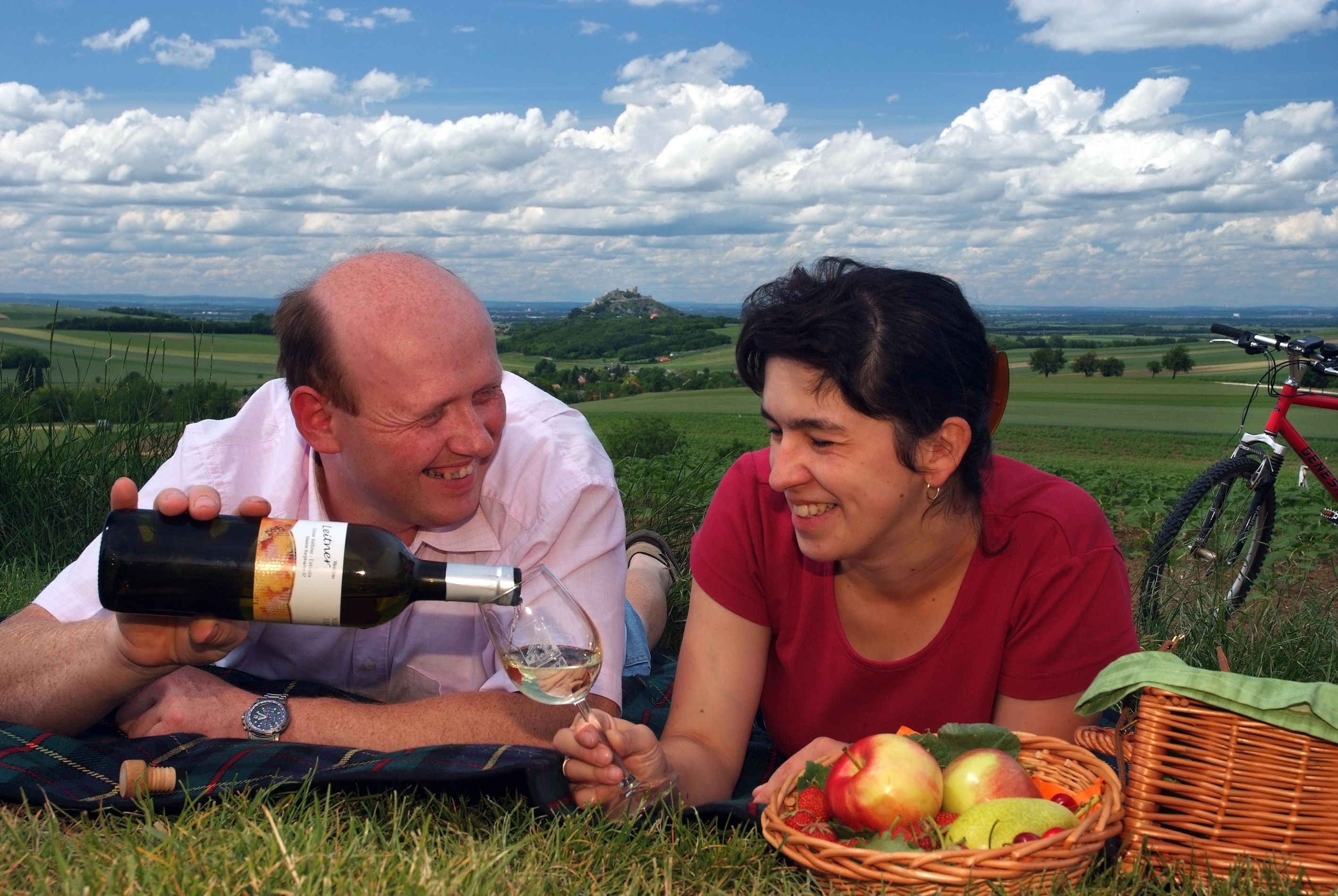 Ein Paar beim Picknick auf einer Wiese mit Wein und Obstkorb, im Hintergrund eine hügelige Landschaft und ein Fahrrad.