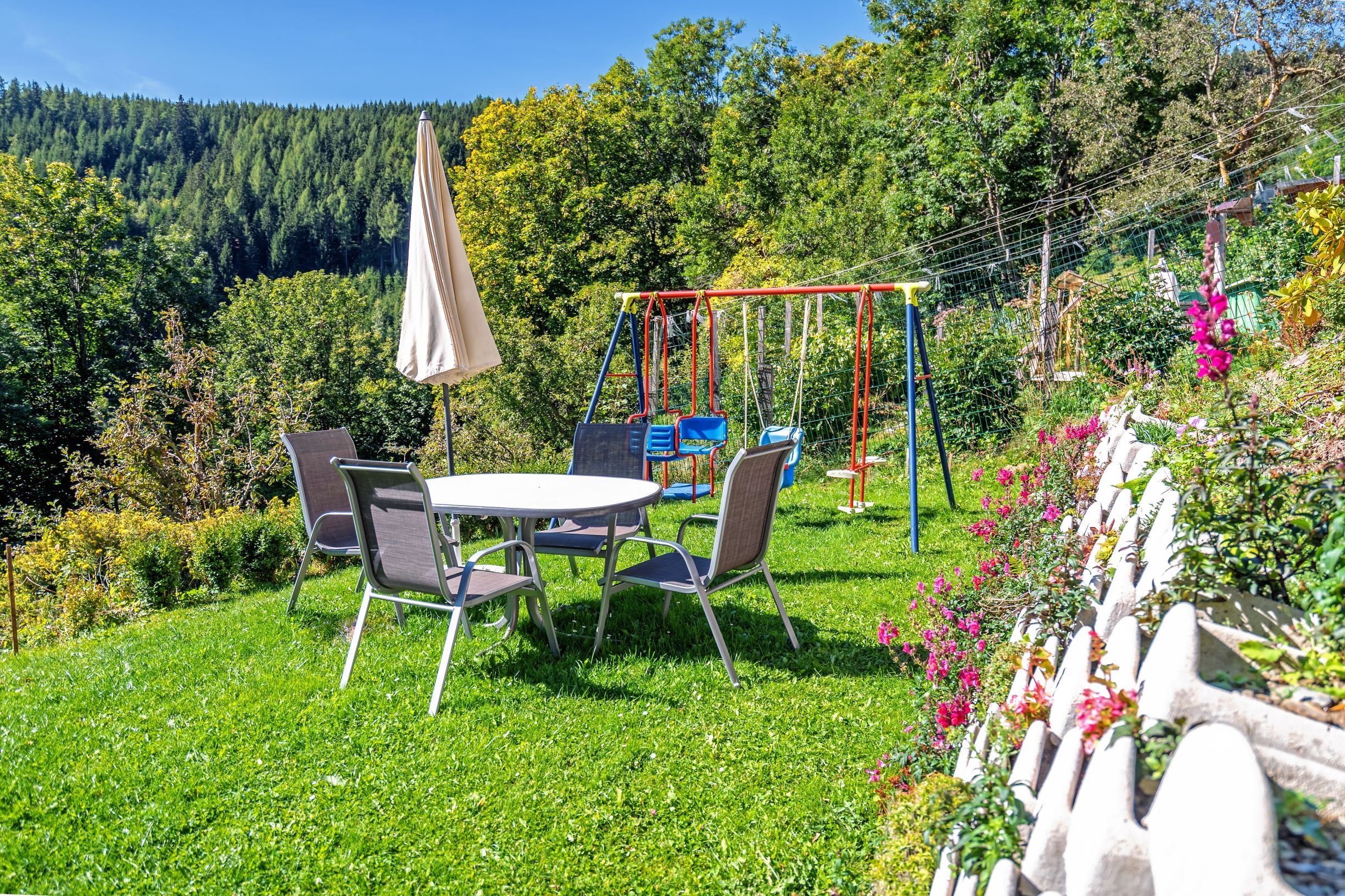 Ein Spielplatz mit Tisch, Stühlen, Sonnenschirm und Schaukeln auf einer Wiese, umgeben von Bäumen und Blumen.