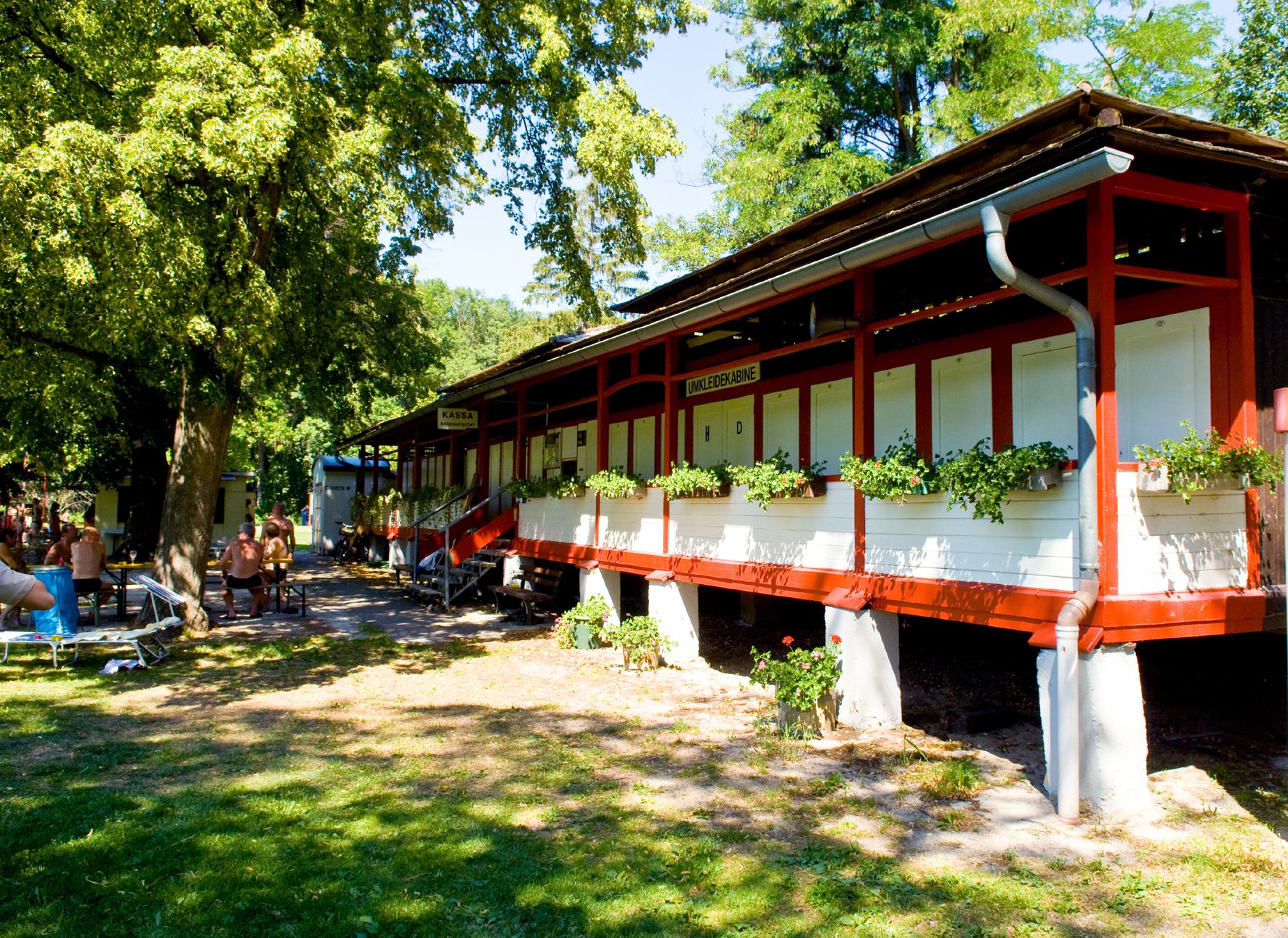 Ein rotes Gebäude mit Umkleidekabinen und Pflanzen davor, umgeben von Bäumen und Menschen auf Bänken.