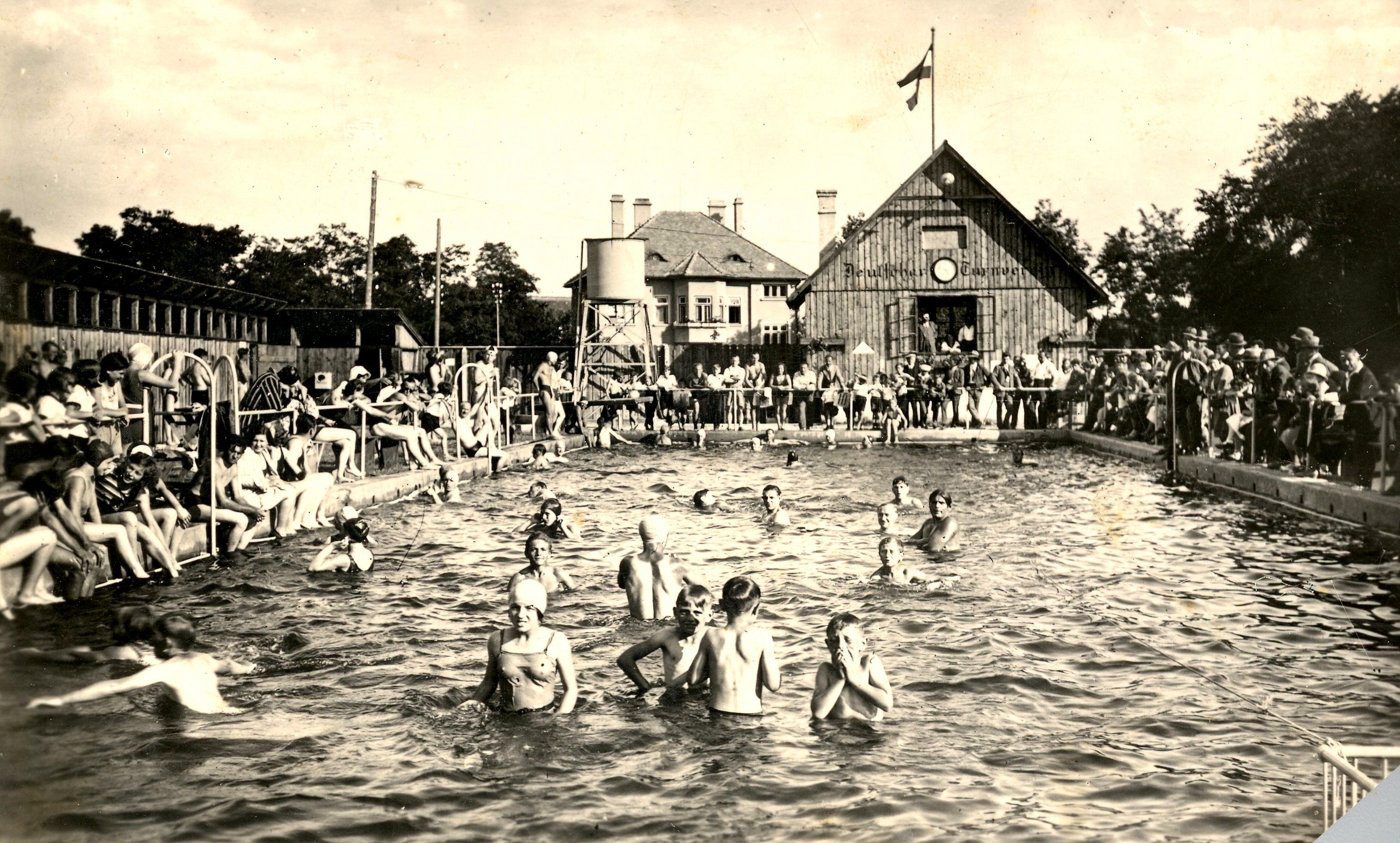Historisches Foto eines Freibads mit vielen Menschen im Wasser und am Beckenrand. Im Hintergrund ein Gebäude mit Fahne.