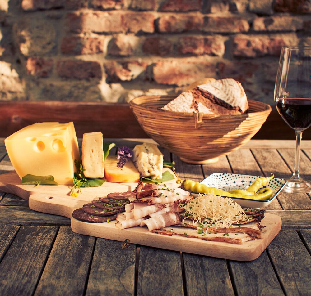 Genießen Sie eine köstliche Auswahl an regionalen Spezialitäten, während die warmen Sonnenstrahlen auf die rustikale Holzplatte scheinen. Ein Glas tiefroter Wein ergänzt die herzhaften Käsesorten und feinen Wurstwaren, die perfekt für einen entspannten Nachmittag im Freien sind.
