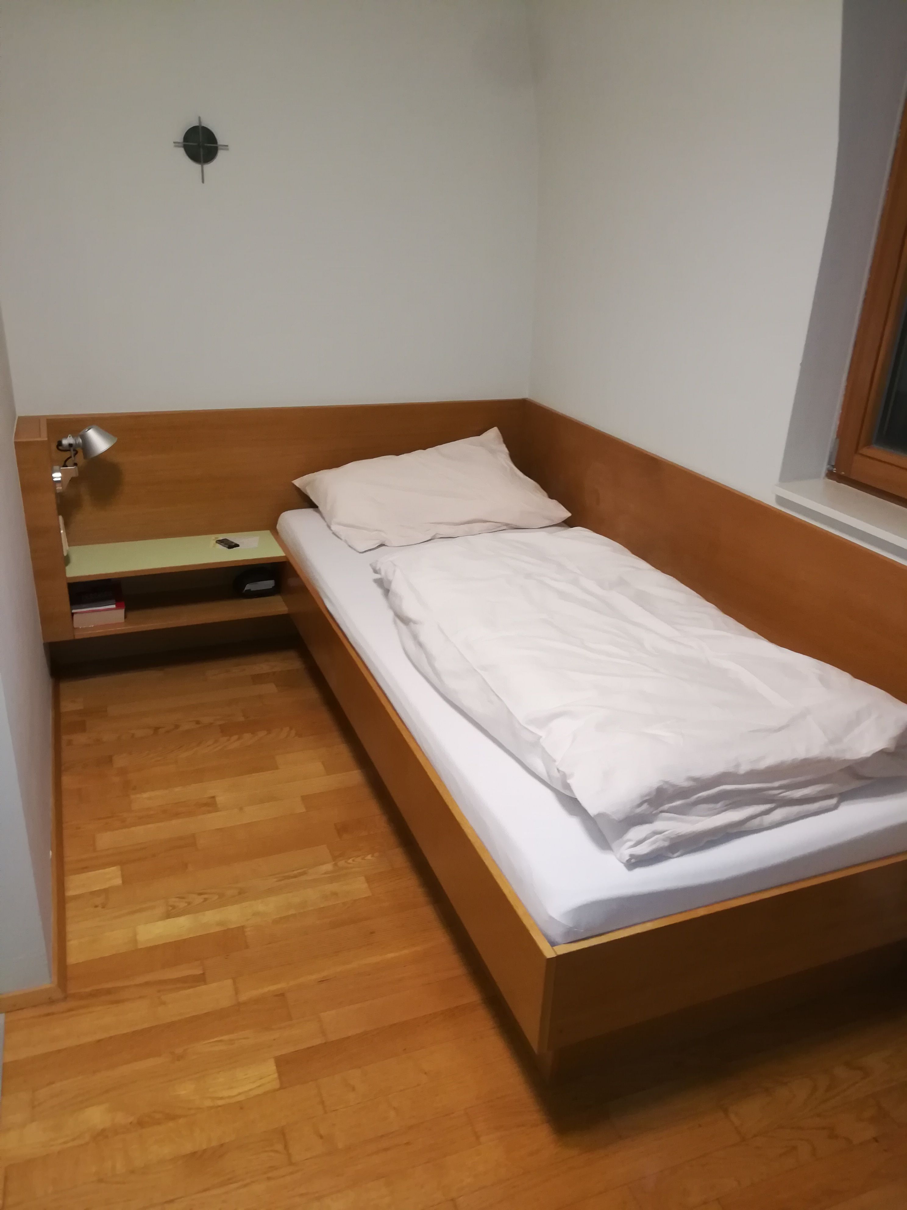 Einzelzimmer mit Holzbett, Nachttisch und Lampe.