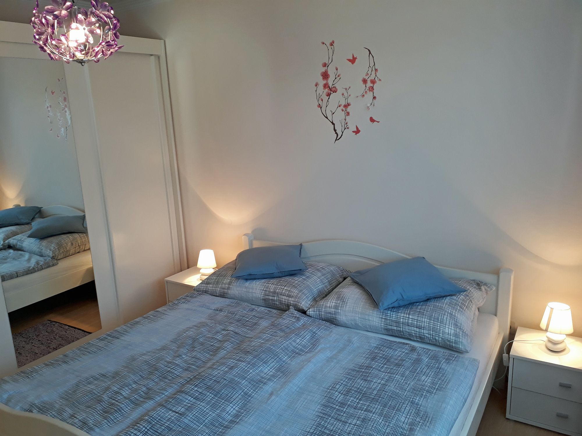 Gemütliches Schlafzimmer mit Doppelbett, blauer Bettwäsche, Nachttischen mit Lampen und einem Wandtattoo mit Blumen und Vögeln.