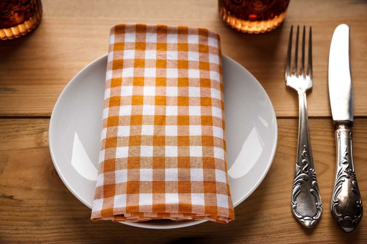 Ein gedeckter Tisch mit einer orange-weiß karierten Serviette auf einem Teller und Besteck daneben.