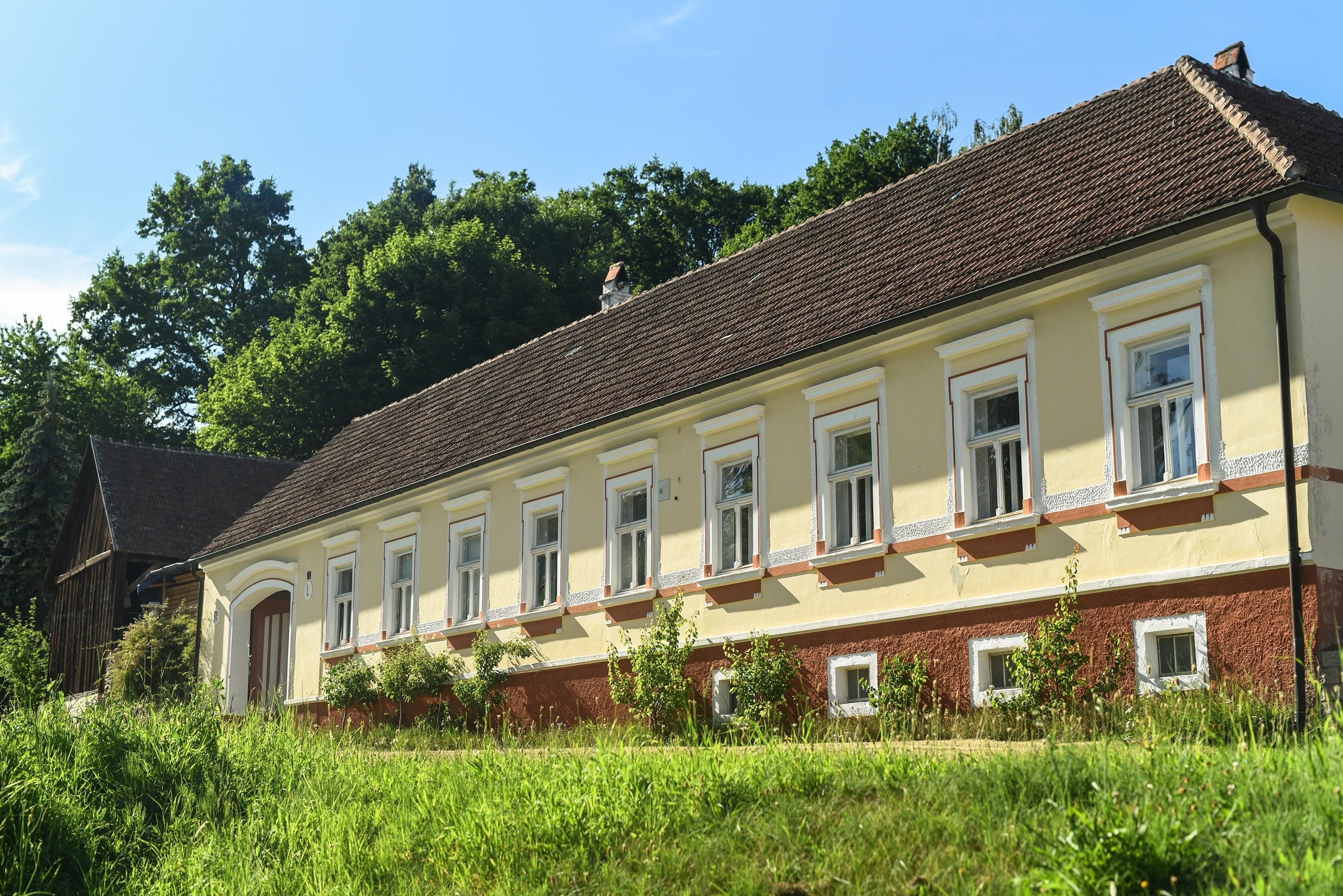 Ein langes, gelbes Landhaus mit roten Dachziegeln und weißen Fensterrahmen, umgeben von grüner Vegetation und Bäumen im Hintergrund.