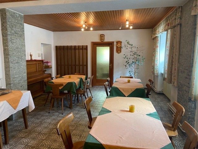 Ein gemütlicher Frühstücksraum mit Holztischen, Stühlen und einem Klavier. Die Tische sind mit Tischdecken gedeckt, und es gibt Fenster mit Vorhängen.