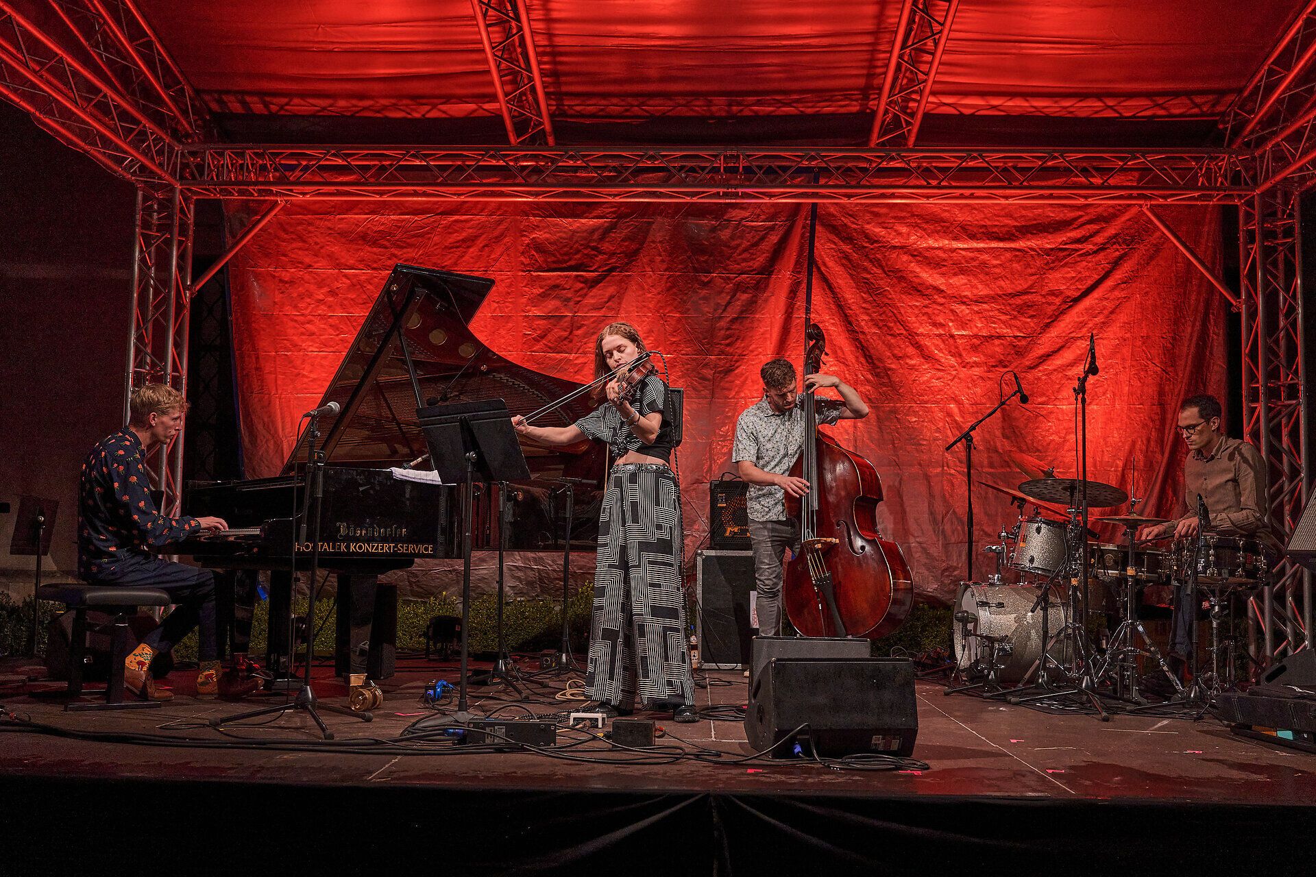 Jazzquartett bei einem Open-Air-Konzert: Pianist am Flügel, Violinistin im Zentrum, Kontrabassist und Schlagzeuger auf einer rot beleuchteten Bühne auf einer Bühne.