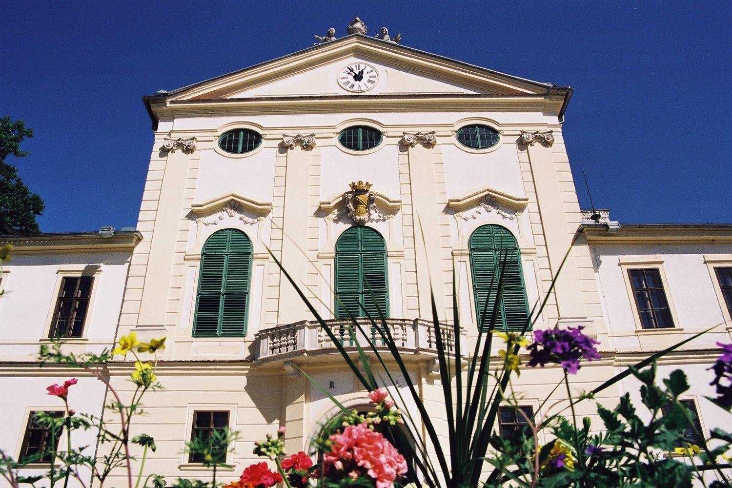 Historisches Gebäude mit grünen Fensterläden und Uhr auf der Fassade, umgeben von bunten Blumen.