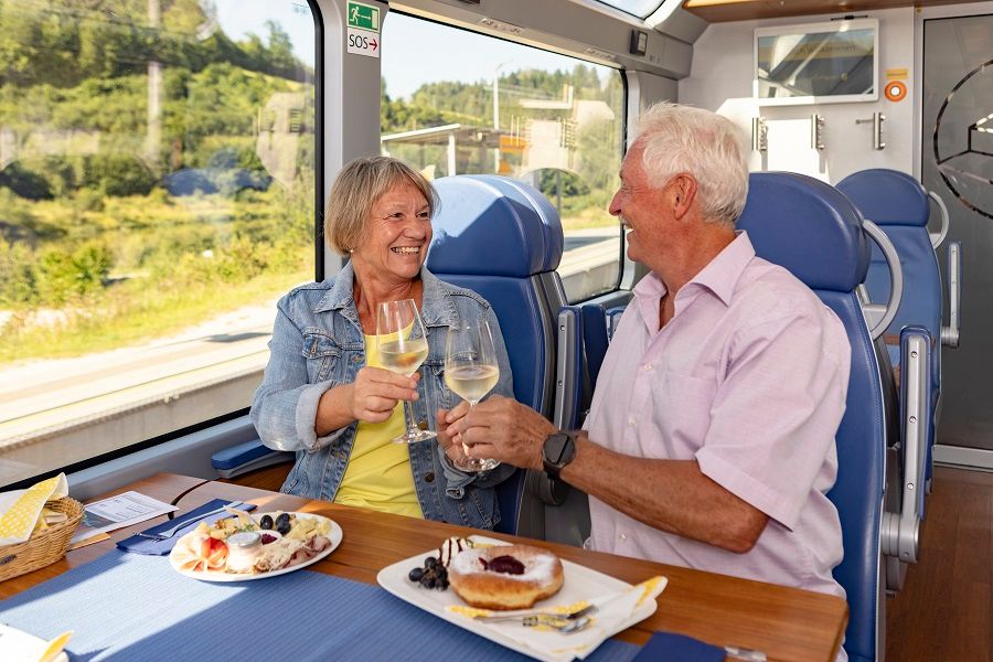 Ein älteres Paar stößt mit Weingläsern in einem Zug an, mit Essen auf dem Tisch.