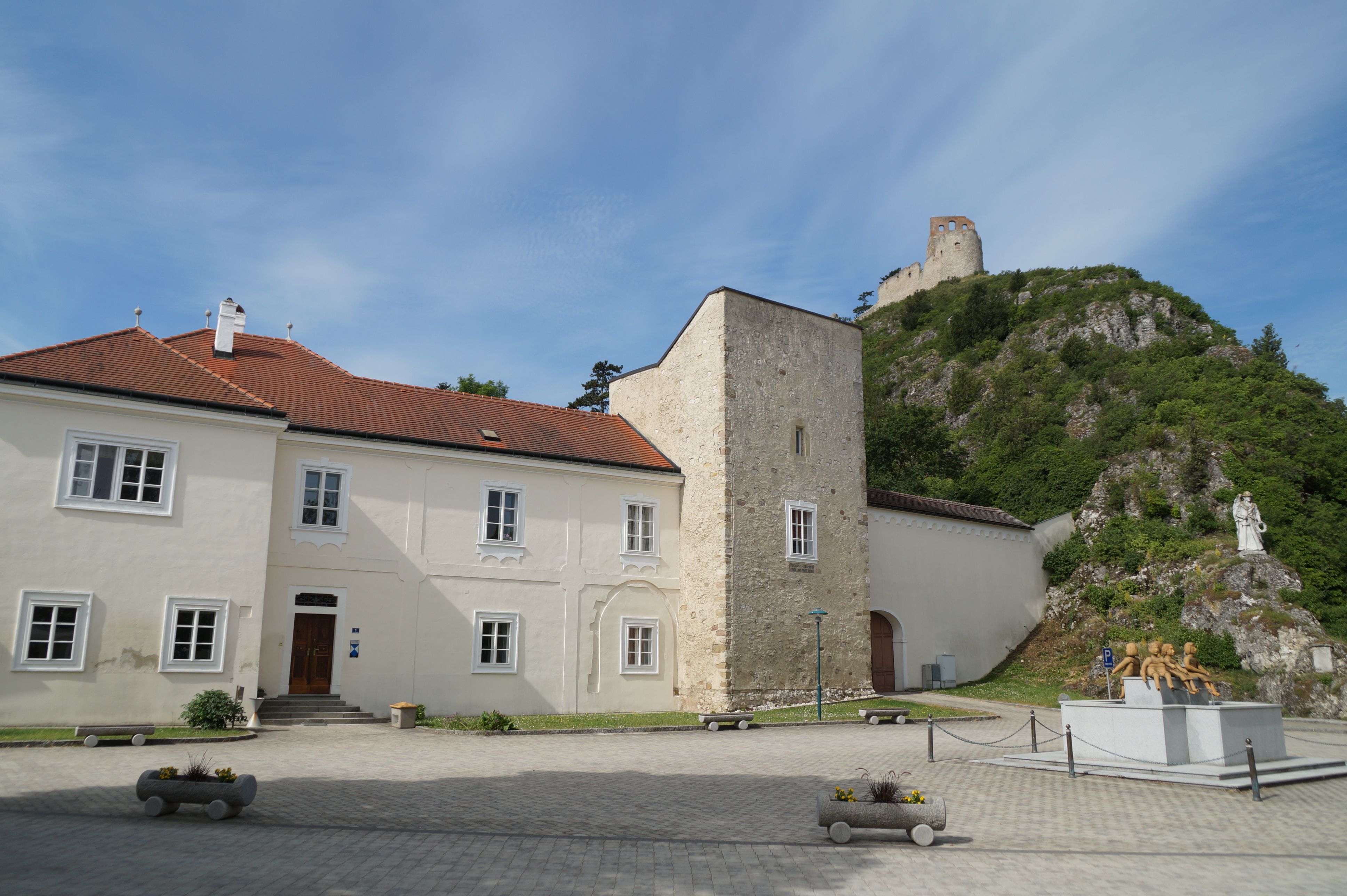 Ein historisches Gebäude mit rotem Ziegeldach und einem steinernen Turm, im Hintergrund eine Burgruine auf einem Hügel.