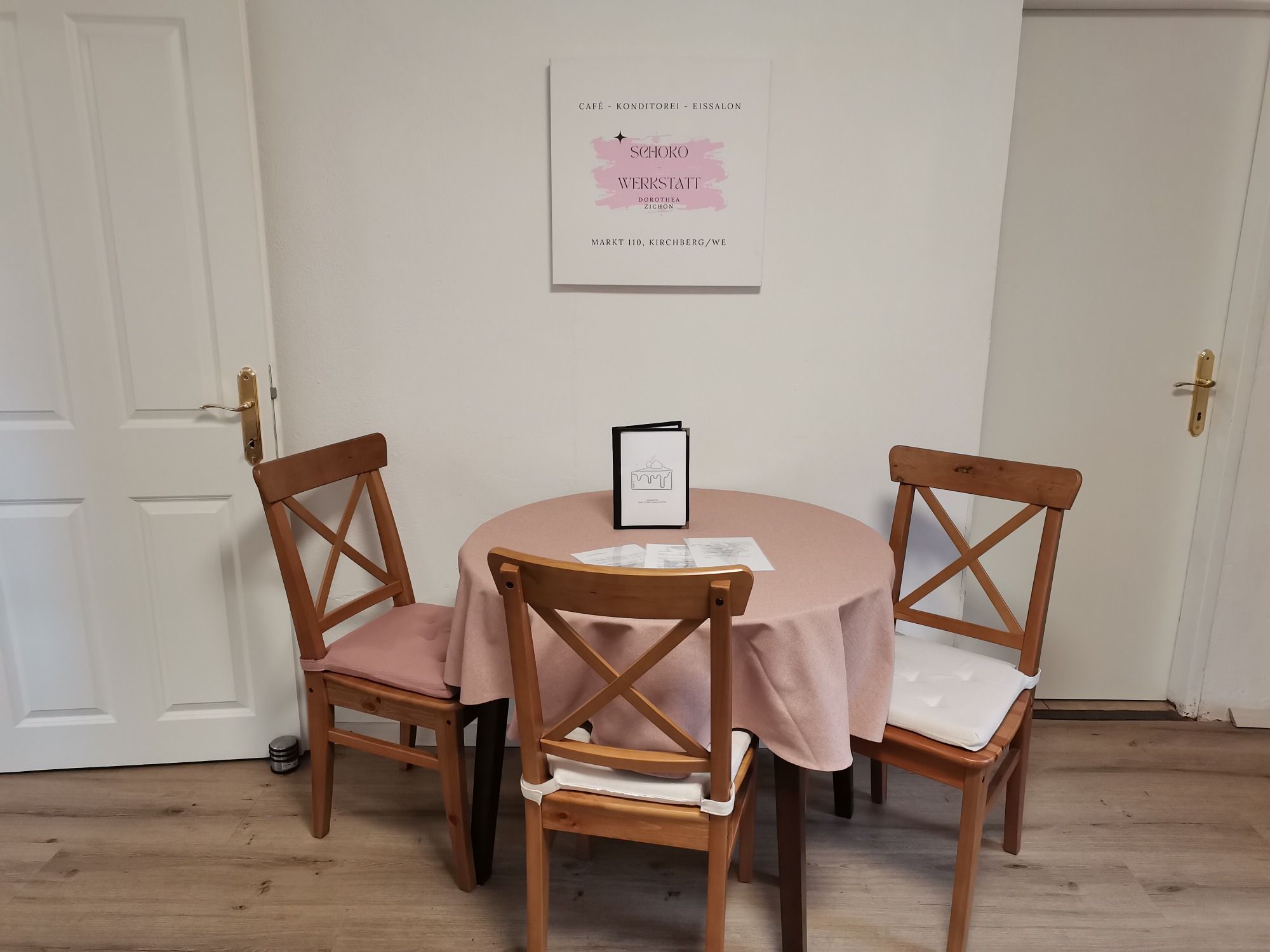 Ein Tisch mit rosa Tischdecke und drei Holzstühlen in einem Café, mit einem Schild an der Wand.