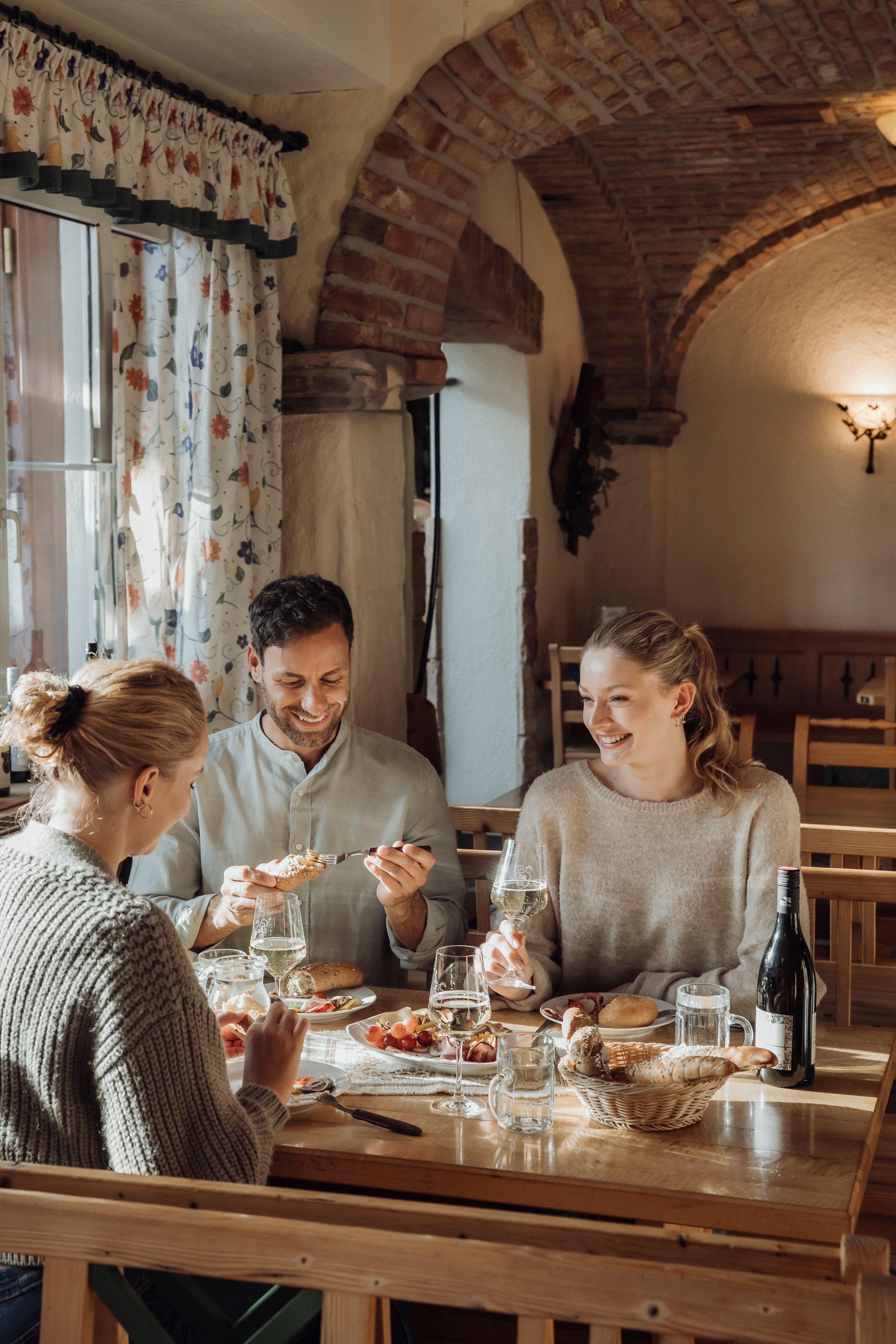 Drei Personen sitzen an einem Tisch in einem gemütlichen Restaurant und genießen Wein und Essen.