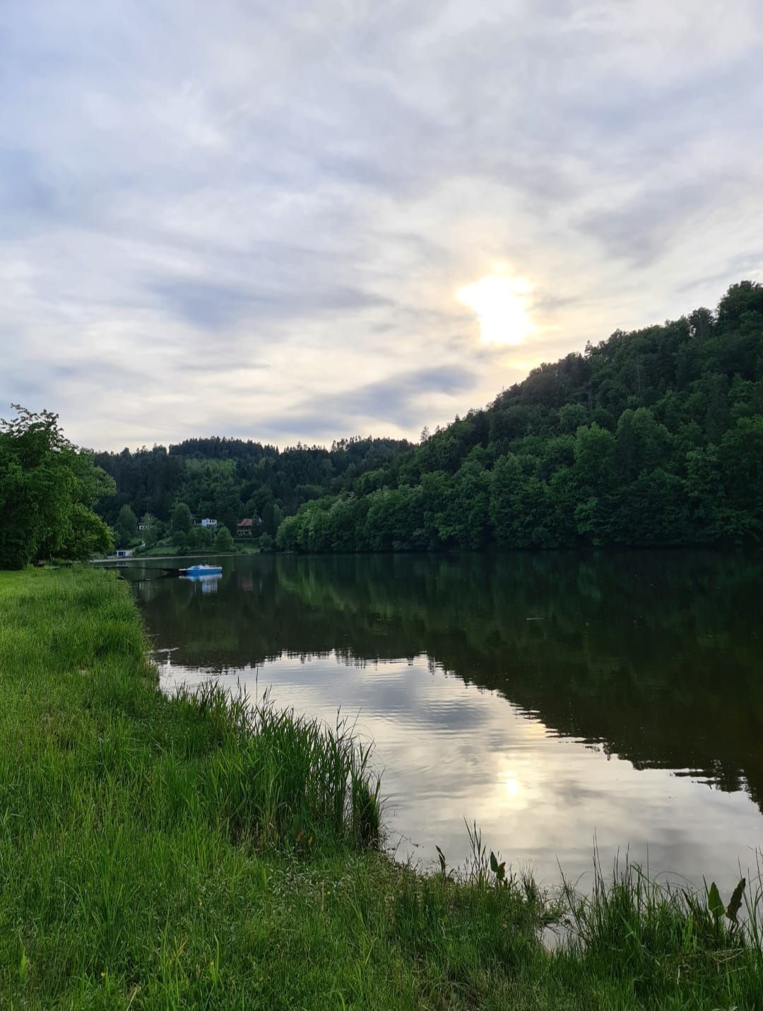 Ein ruhiger See mit bewaldeten Hügeln im Hintergrund und einer leicht bewölkten Abendsonne.