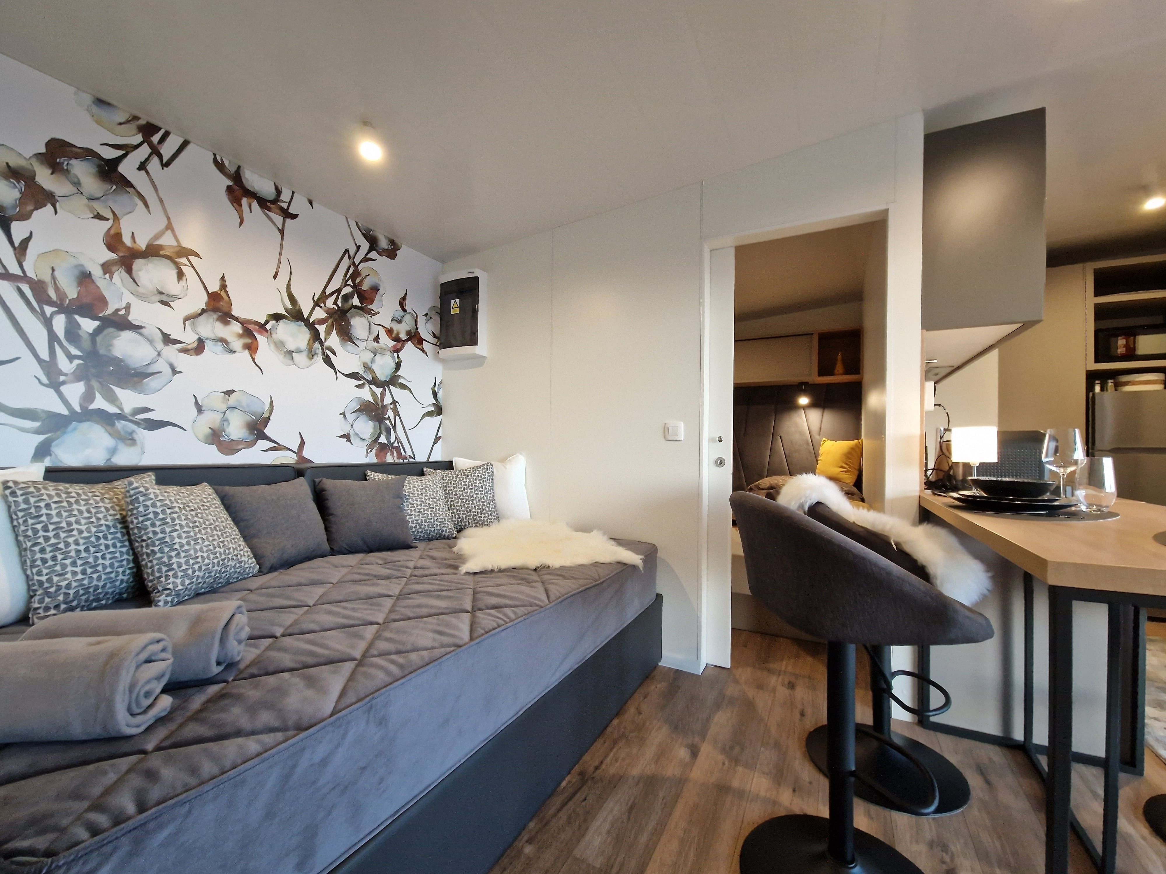 Innenansicht eines modernen Chalets mit grauem Sofa, dekorativer Blumenwand, Barhocker und Esstisch.