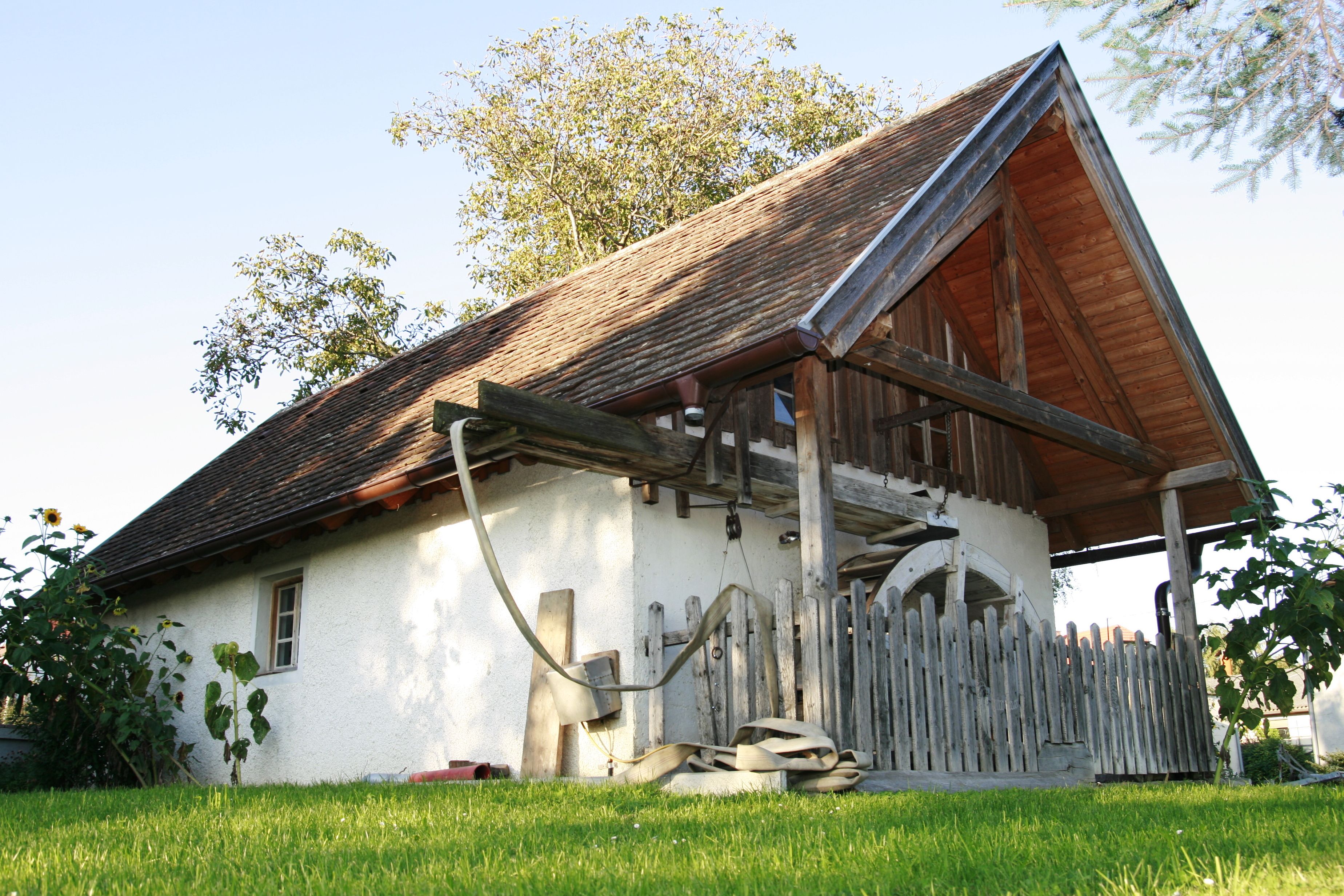 Traditionelles Gebäude mit Holzdach und Zaun, umgeben von grüner Wiese und Bäumen.