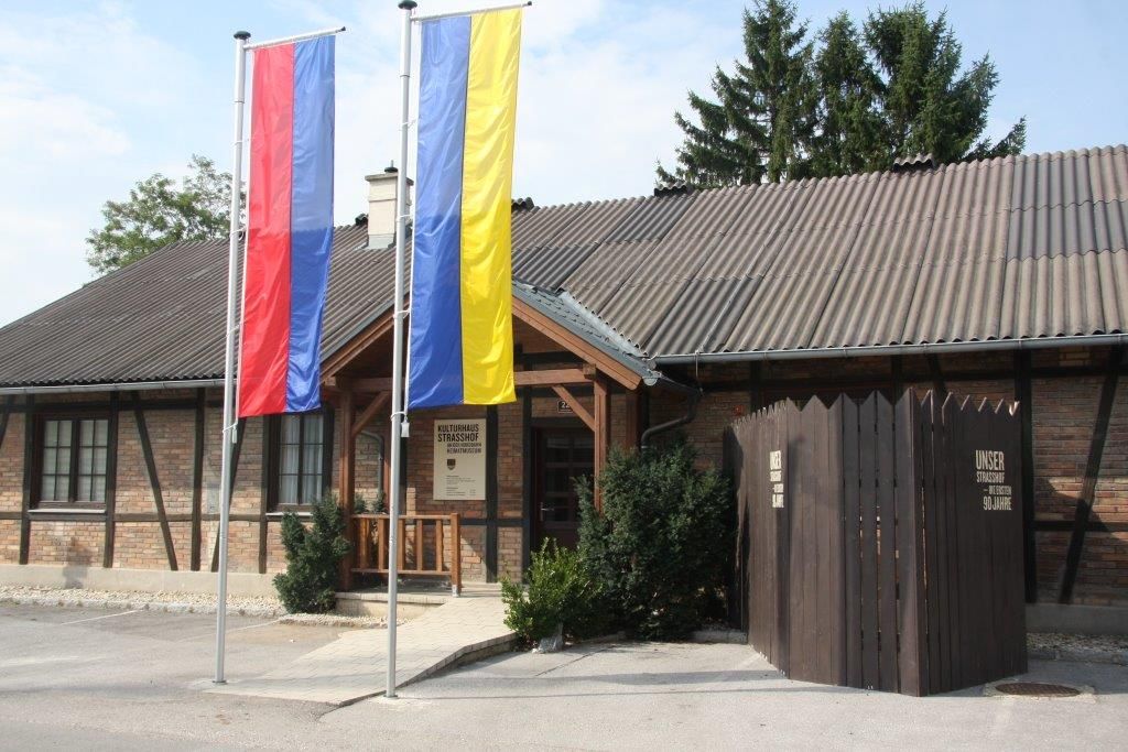 Eingang des Heimatmuseums Strasshof mit zwei Fahnenmasten und einem Holzzaun.