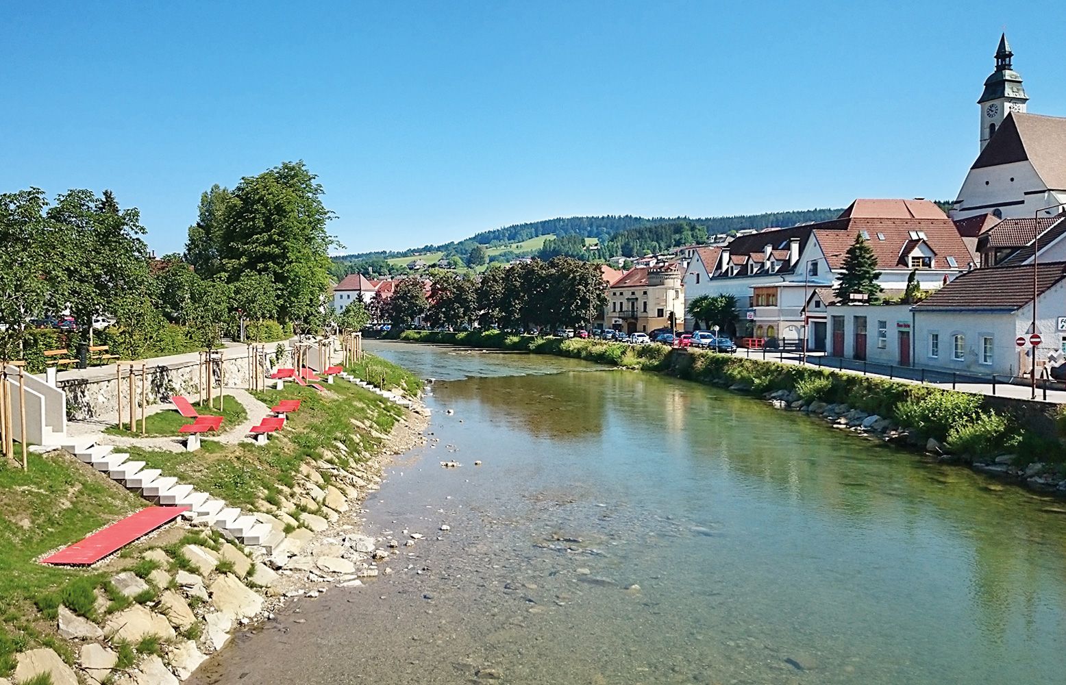 Fluss mit Uferpromenade und roten Liegen, Stadt im Hintergrund.