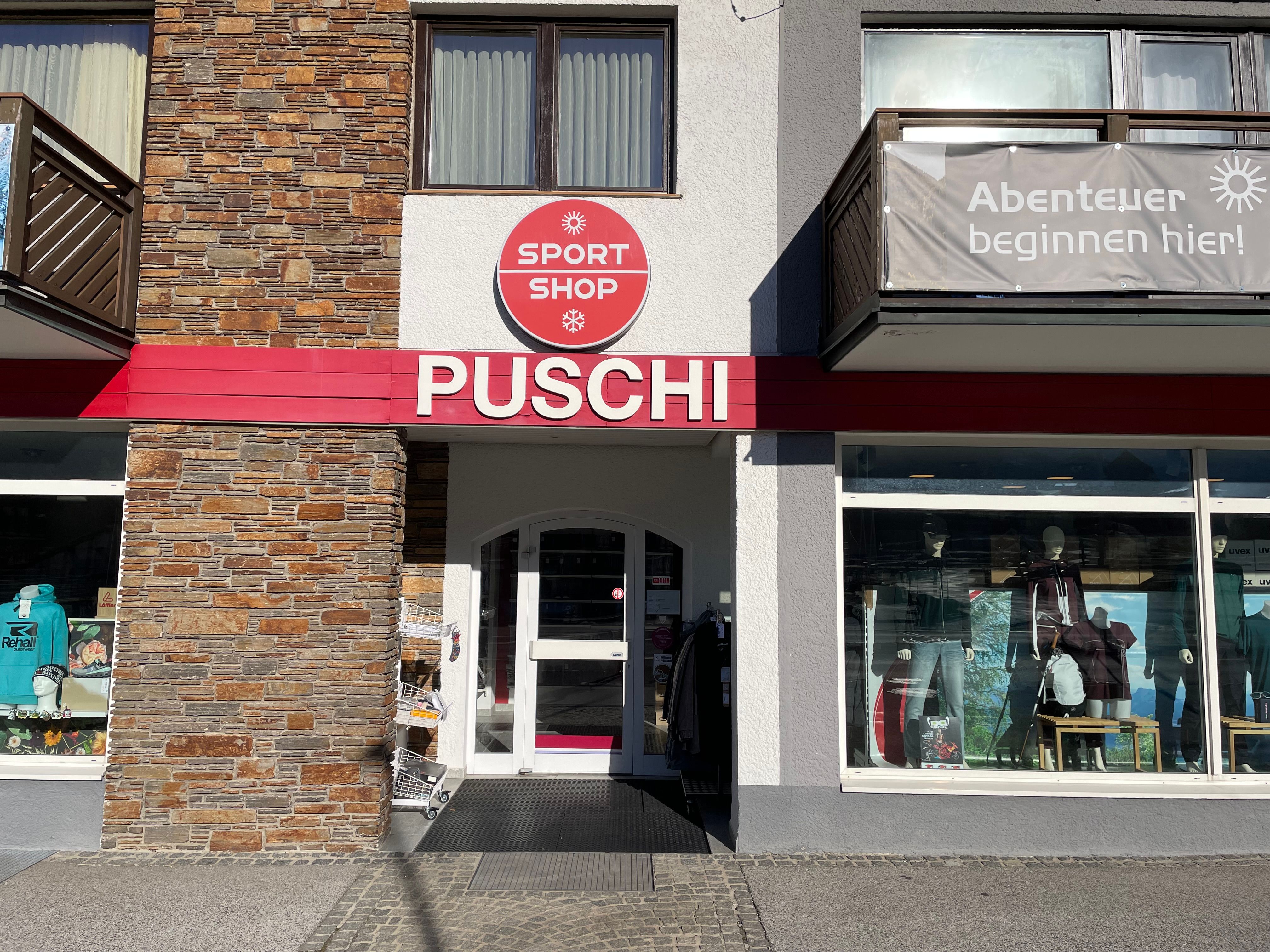 Eingangsbereich eines Sportgeschäfts mit der Aufschrift 'PUSCHI' und einem Schild 'SPORT SHOP'. Schaufenster mit Sportbekleidung und Ausrüstung.