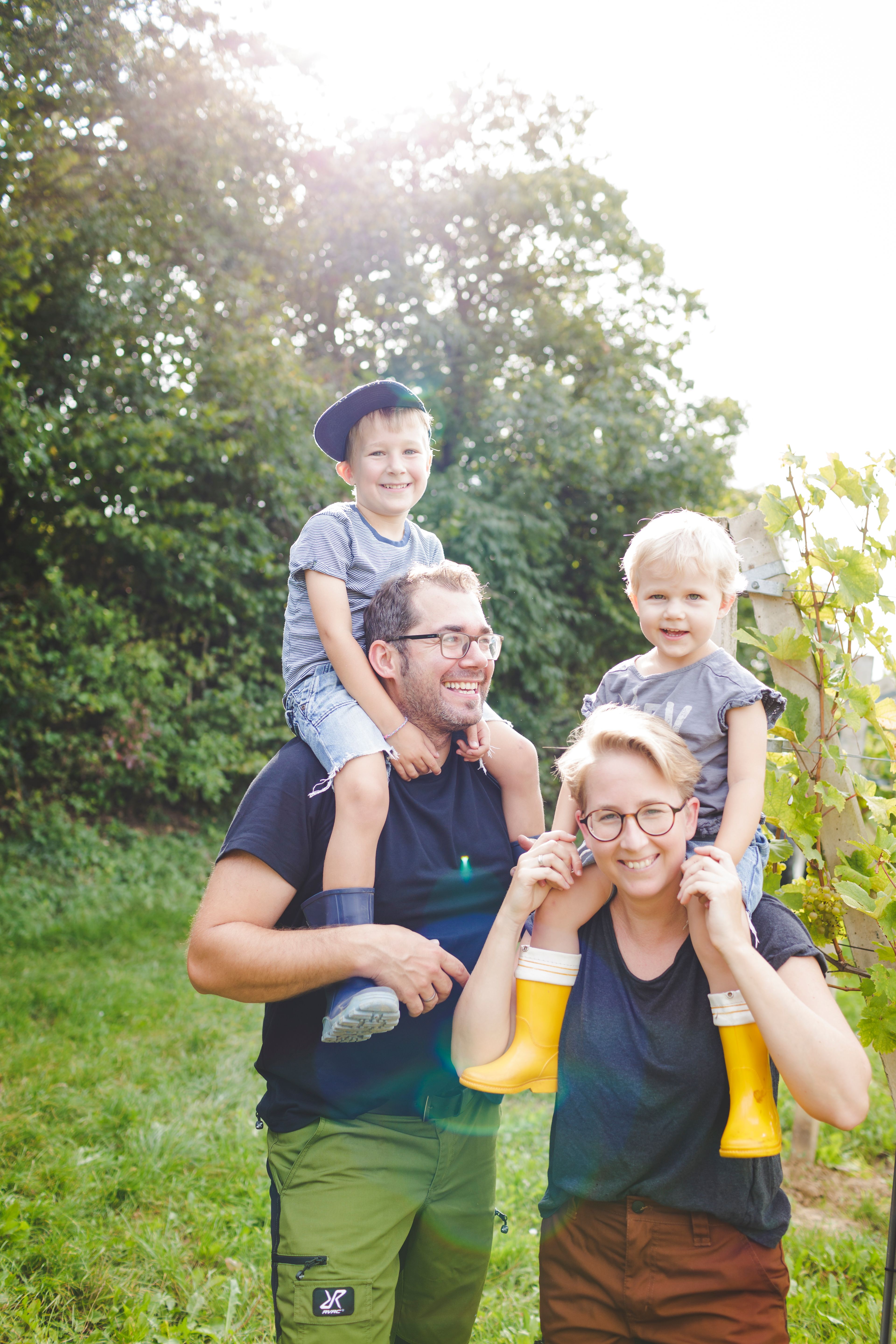 Familie im Freien, zwei Erwachsene tragen Kinder auf den Schultern, sonniger Tag.