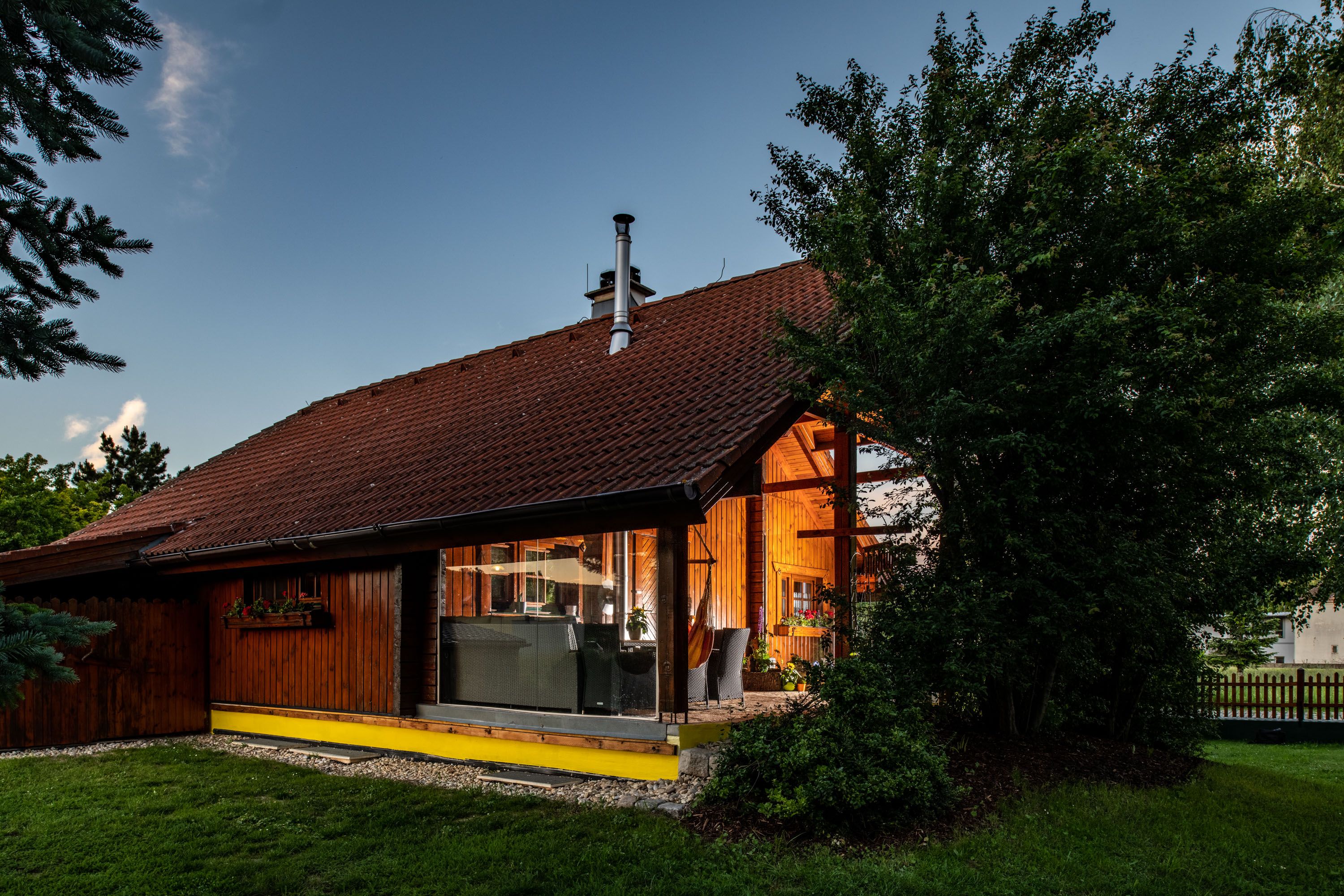 Außenansicht eines Holzhauses mit großen Fenstern und Garten, umgeben von Bäumen.