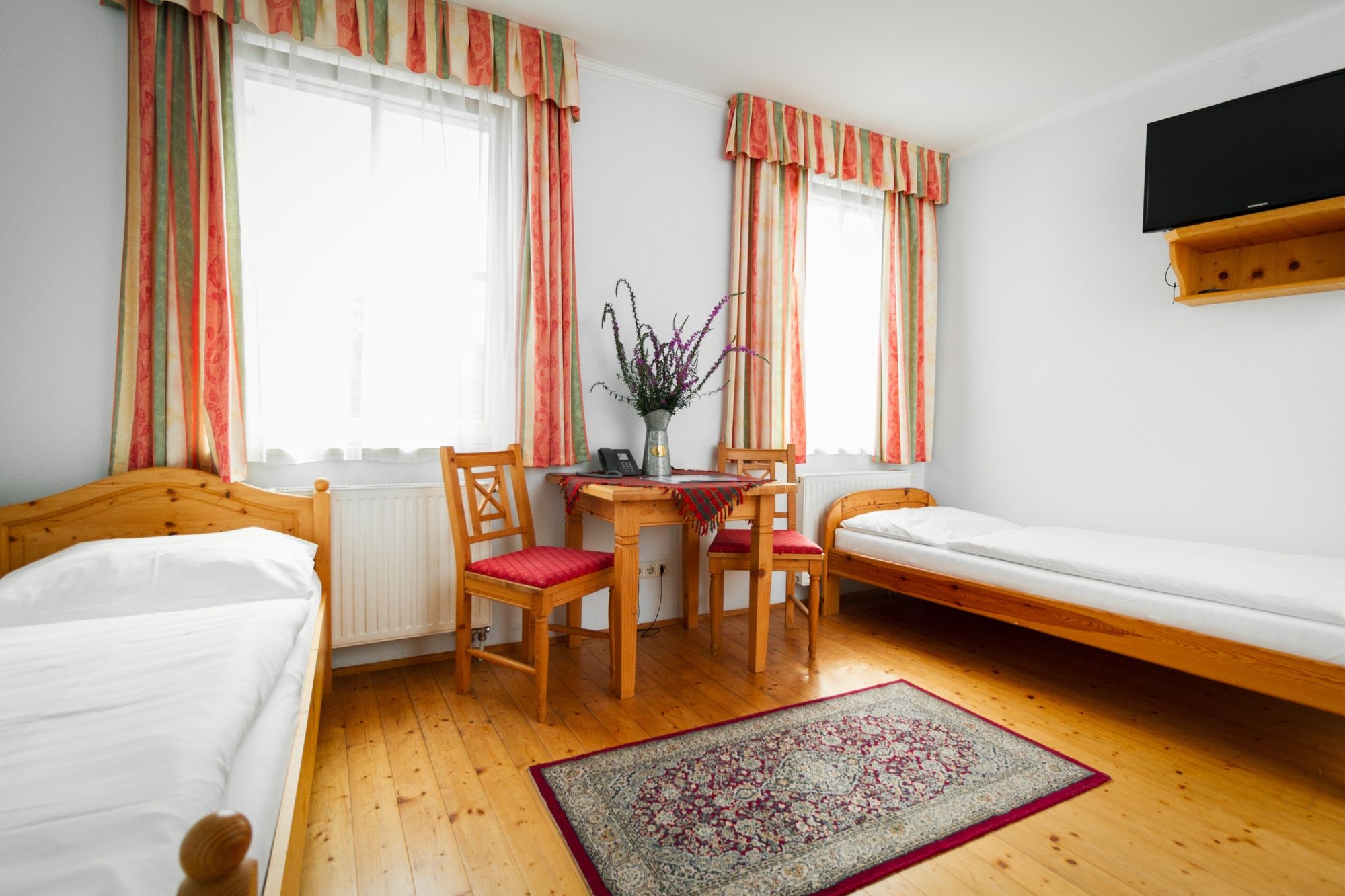 Ein einfach eingerichtetes Zimmer mit zwei Einzelbetten, einem Tisch mit zwei Stühlen, einem Teppich und einem Fernseher an der Wand.