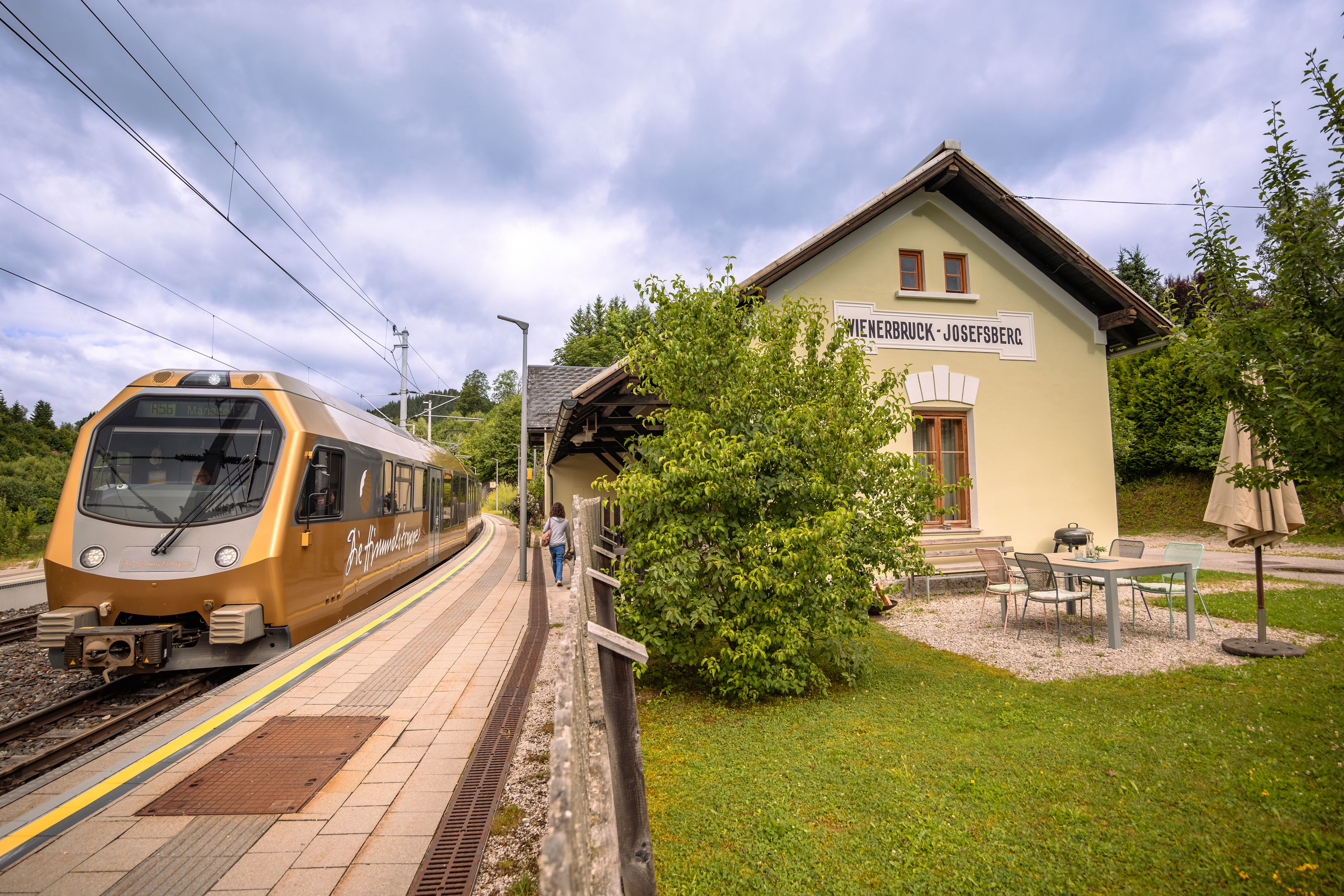 Ein goldener Zug hält am Bahnhof Wienerbruck-Josefsberg, umgeben von grüner Landschaft und einem kleinen Sitzbereich im Freien.
