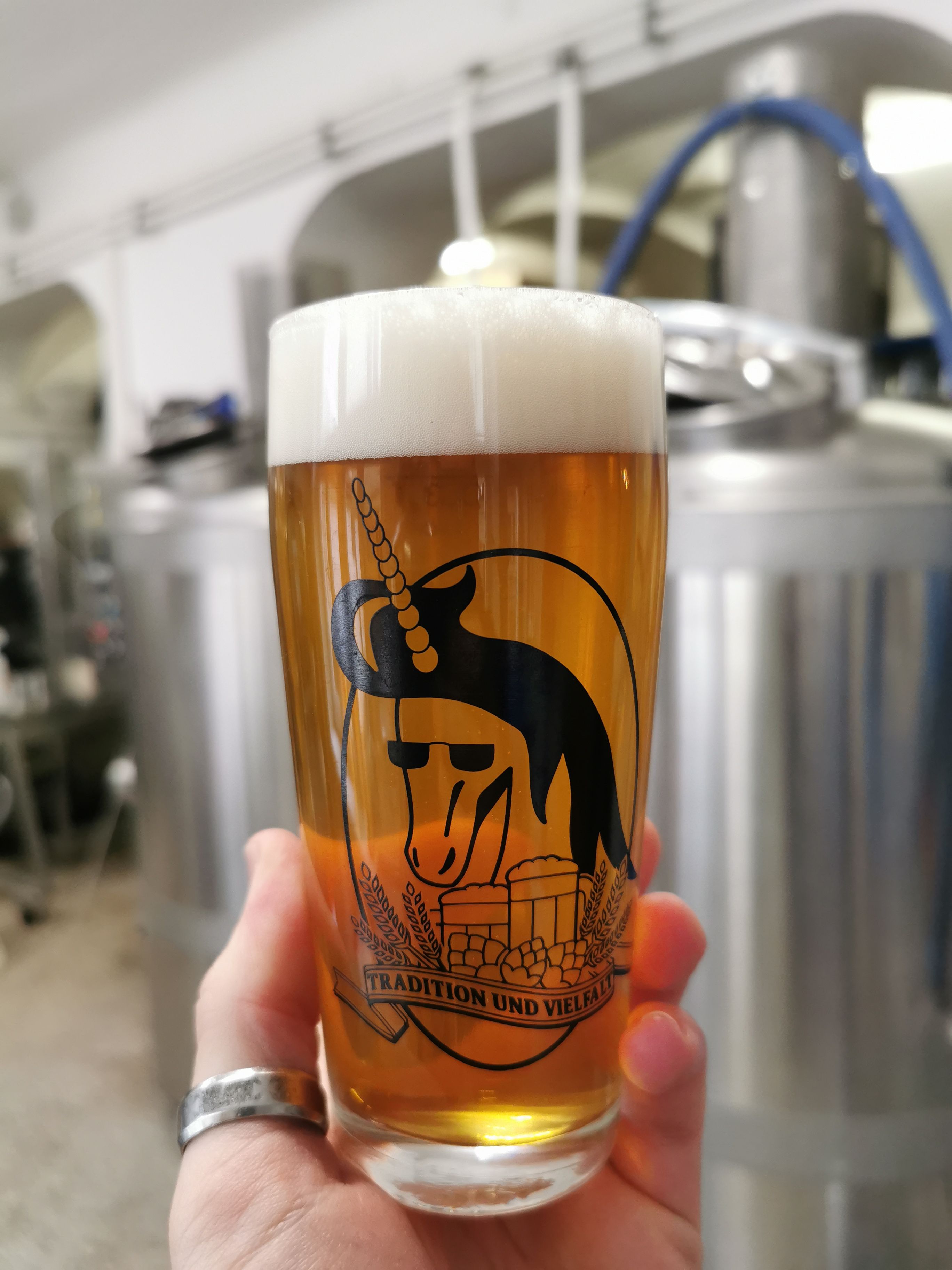 Ein Bierglas mit Einhorn-Logo und der Aufschrift 'Tradition und Vielfalt' vor Brauereiausrüstung.