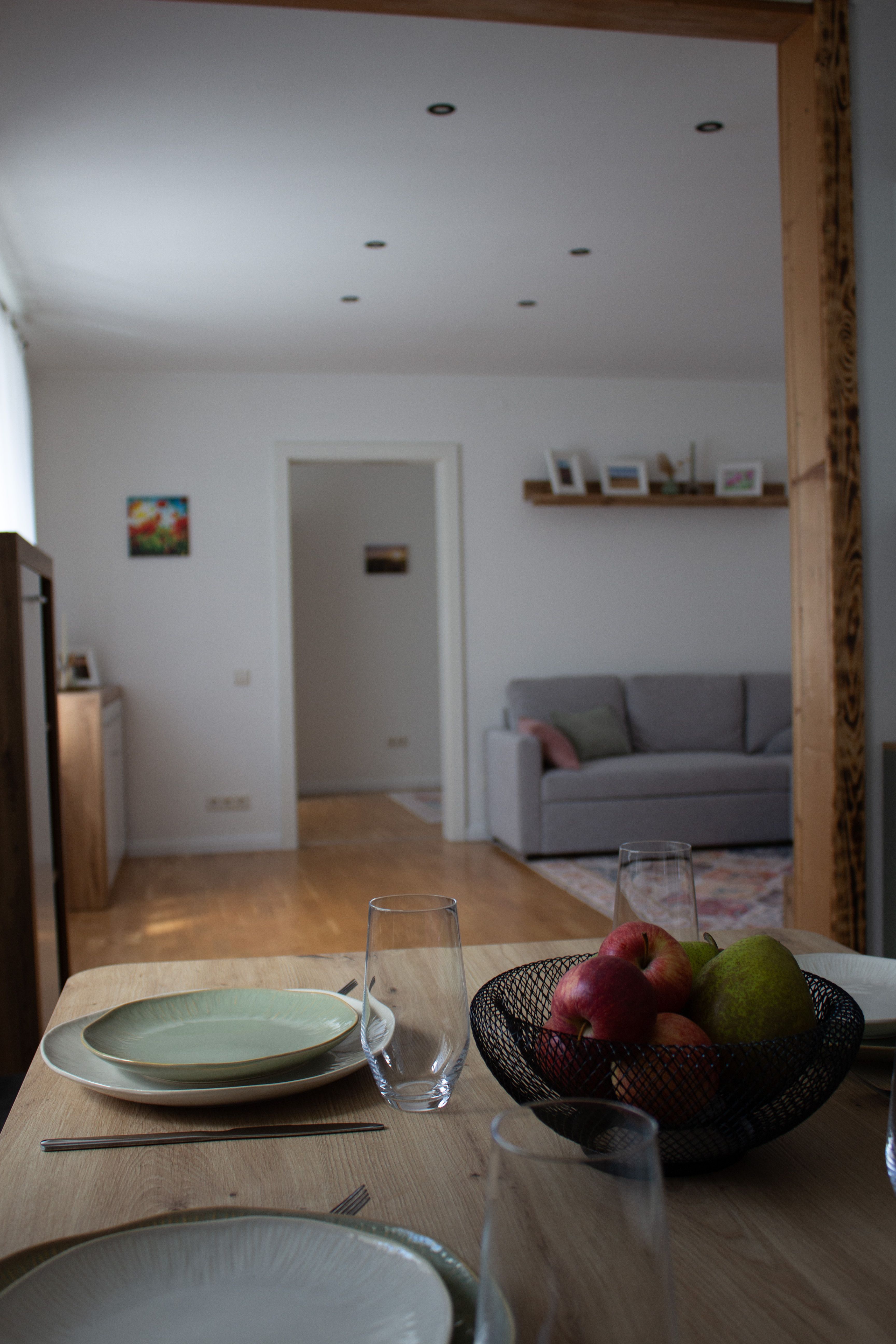 Ein Essbereich mit gedecktem Tisch, Obstschale und Blick auf ein Wohnzimmer mit grauem Sofa.