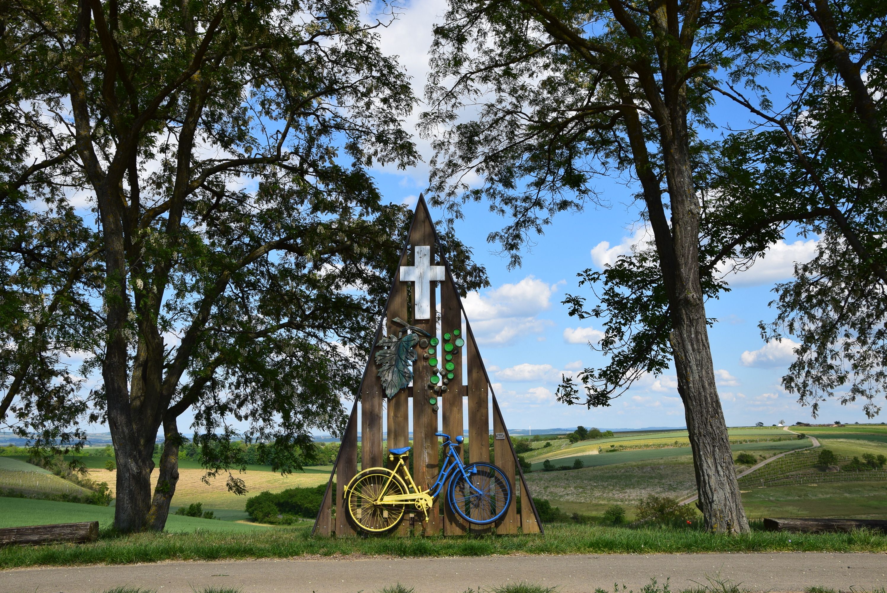 Ein Marterl mit einem gelben und blauen Fahrrad, umgeben von Bäumen und Feldern unter blauem Himmel.