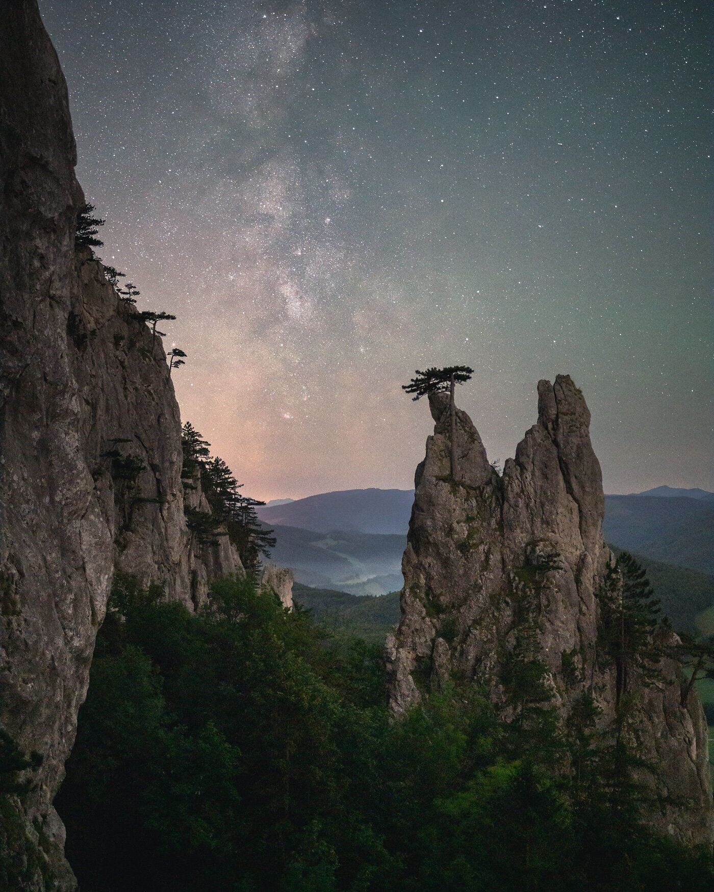Die majestätischen Felsen erheben sich stolz über die üppige Waldlandschaft, während der klare Nachthimmel mit funkelnden Sternen geschmückt ist. Ein einsamer Baum auf einem der Gipfel symbolisiert die unberührte Natur und lädt Wanderer ein, die Ruhe und Schönheit dieser Region zu genießen.