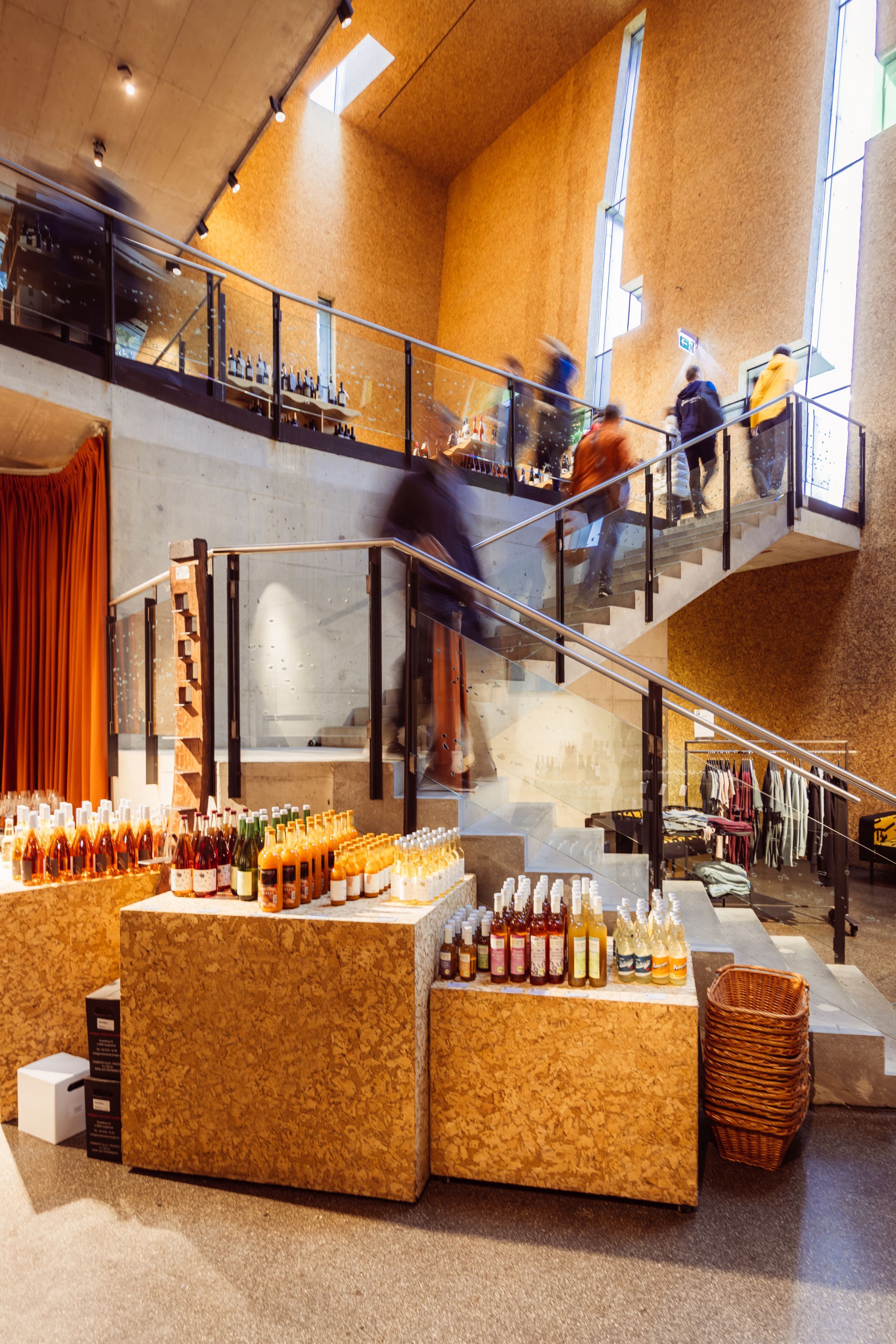 Innenansicht der Steininger Weinwelt mit Shop und Vinothek: Treppe, Glasgeländer, ausgestellte Weinflaschen und mehrere Personen auf der Galerie.