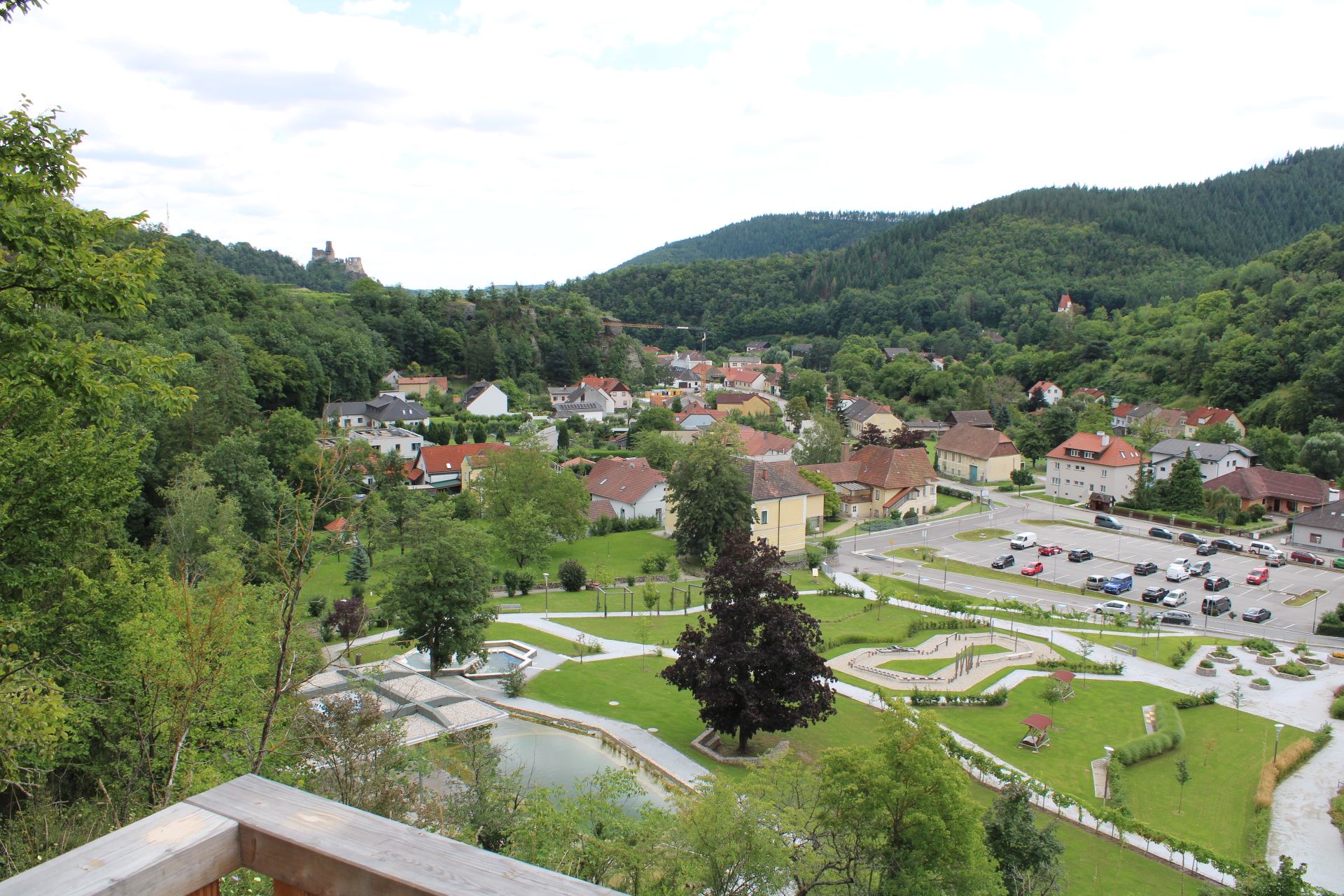 Blick von einer Aussichtsplattform auf eine Stadt mit umliegenden Wäldern und einer Burgruine im Hintergrund.