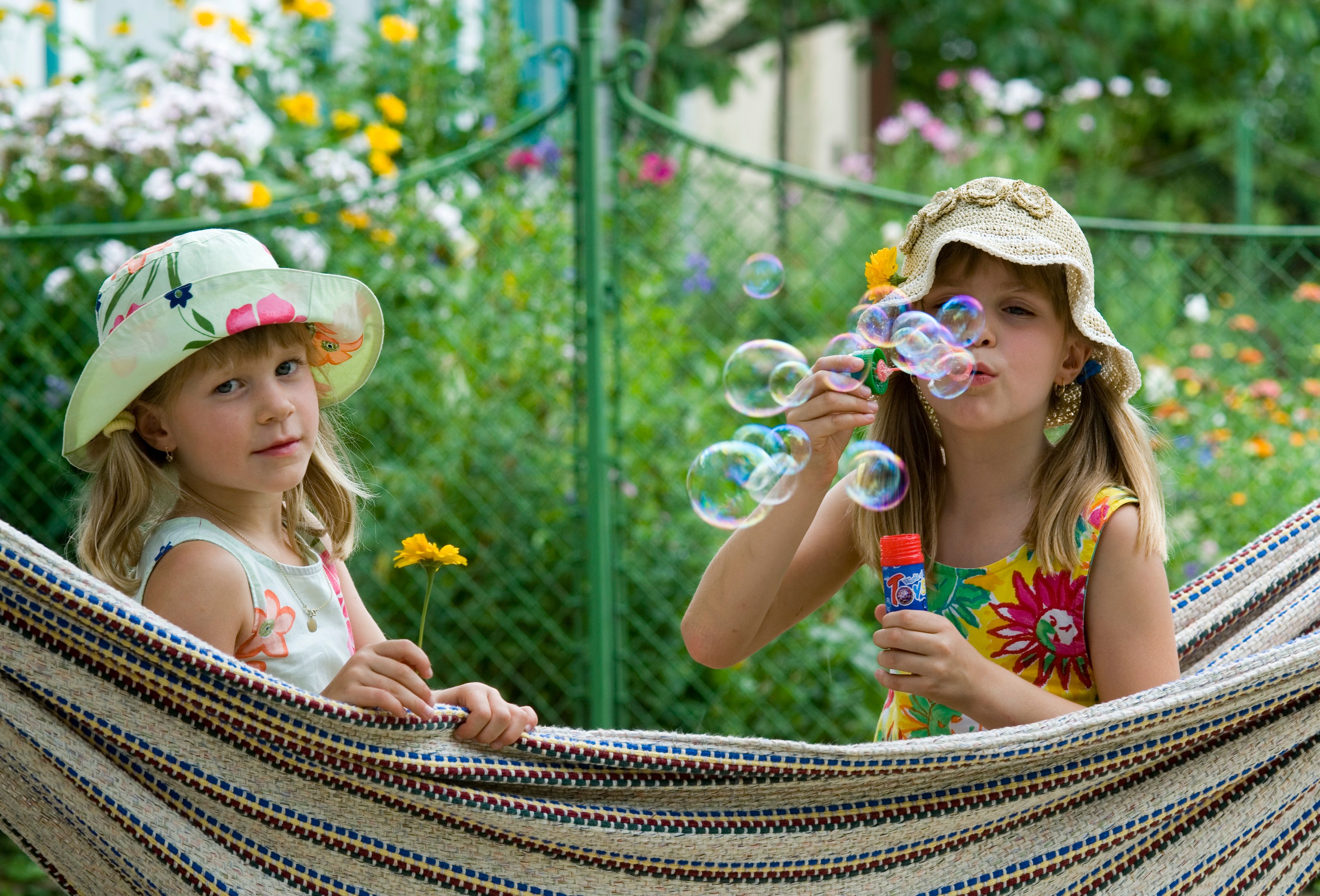 Zwei Mädchen in Sommerkleidern und Hüten sitzen in einer Hängematte. Eines bläst Seifenblasen, das andere hält eine gelbe Blume. Im Hintergrund blühende Blumen.