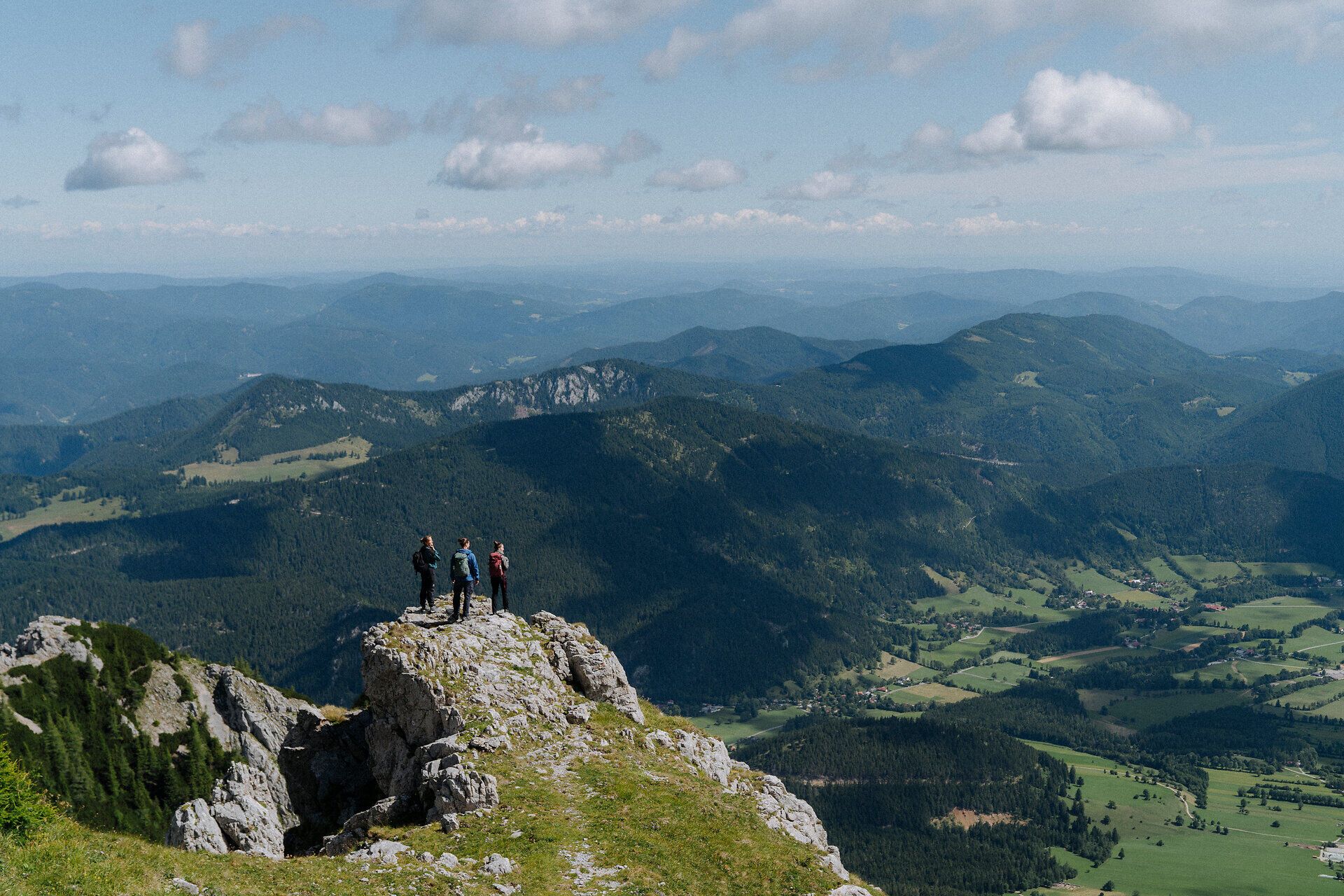 Drei Wandernde erklimmen einen felsigen Gipfel, von dem aus ein beeindruckender Blick über ein weitläufiges Bergpanorama und grüne Täler unter einem blauen Himmel mit weißen Wolken geboten wird, was ein Gefühl von Freiheit und Abenteuer vermittelt.