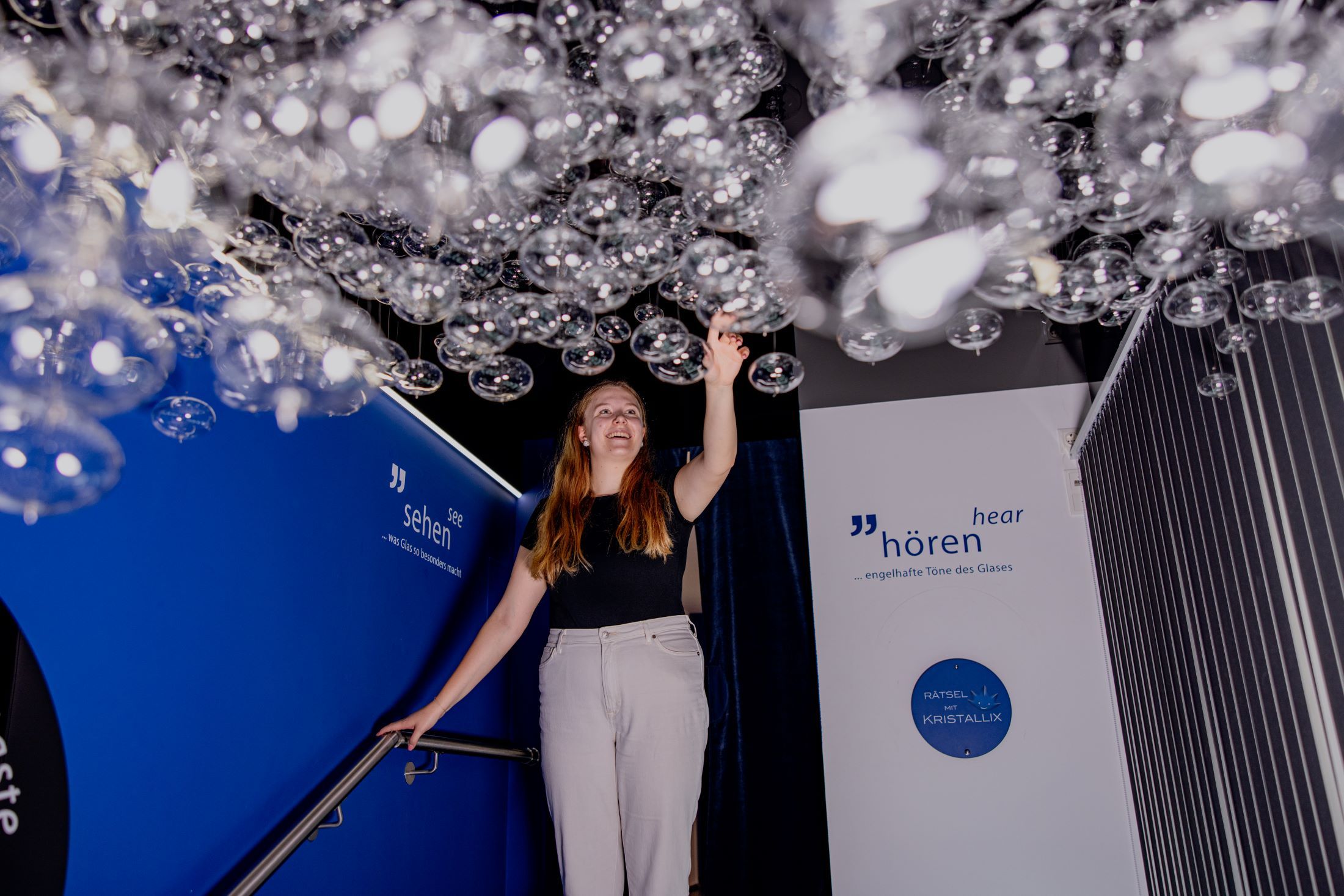 Eine Frau berührt hängende Glasblasen in einer Ausstellung mit blauen Wänden und Beschriftungen.
