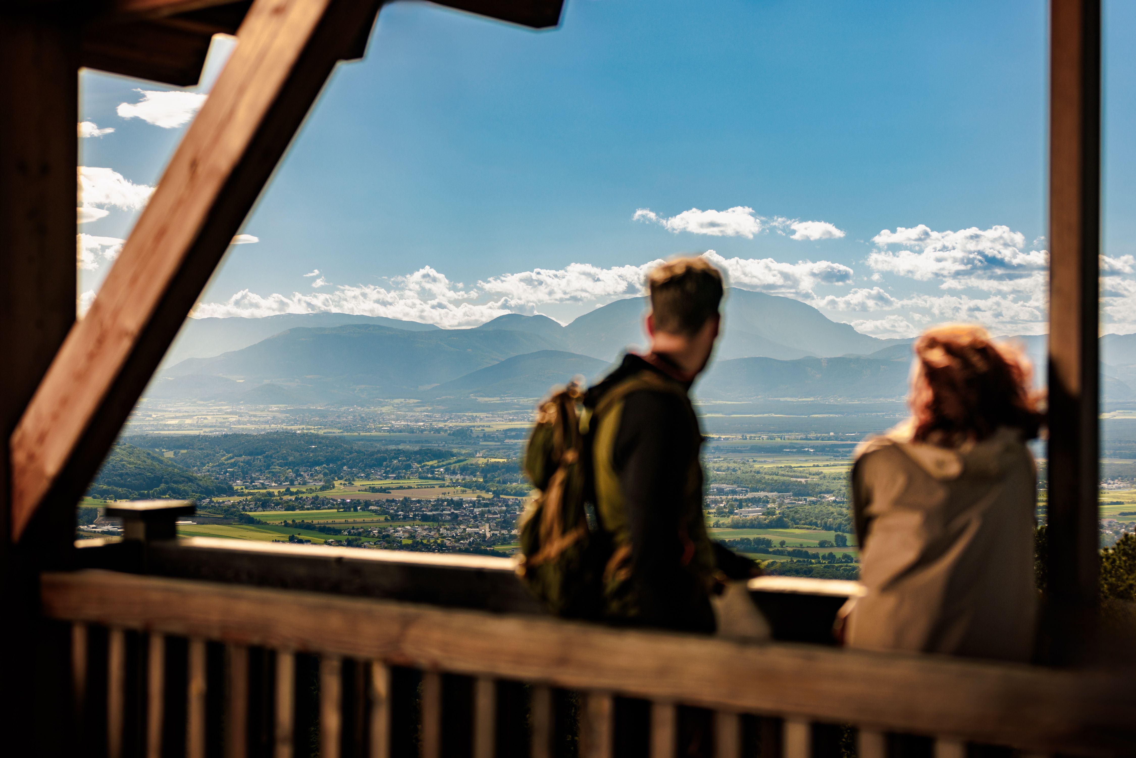 Zwei Personen genießen die Aussicht von einem Aussichtspunkt auf eine weite Landschaft mit Bergen im Hintergrund.