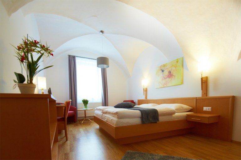 Helles Hotelzimmer mit Doppelbett, Schreibtisch, Pflanzen und Kunstwerk an der Wand.