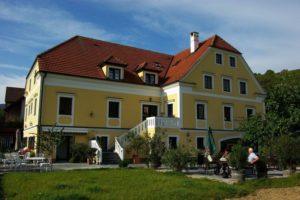 Gelbes Gebäude mit rotem Dach und Terrasse, umgeben von Bäumen.