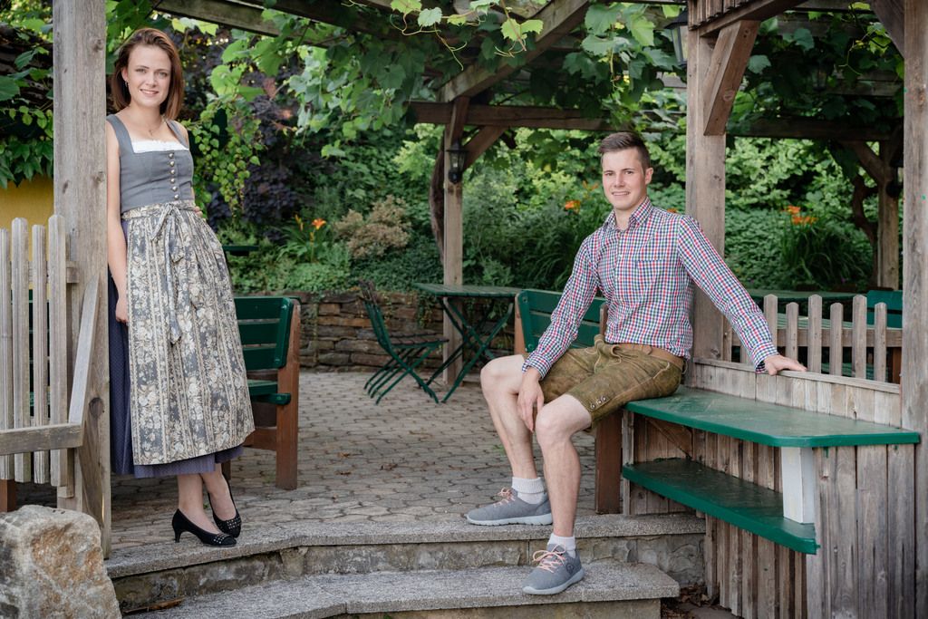 Ein Mann in Lederhose und eine Frau im Dirndl posieren in einem Garten mit Holzkonstruktionen.