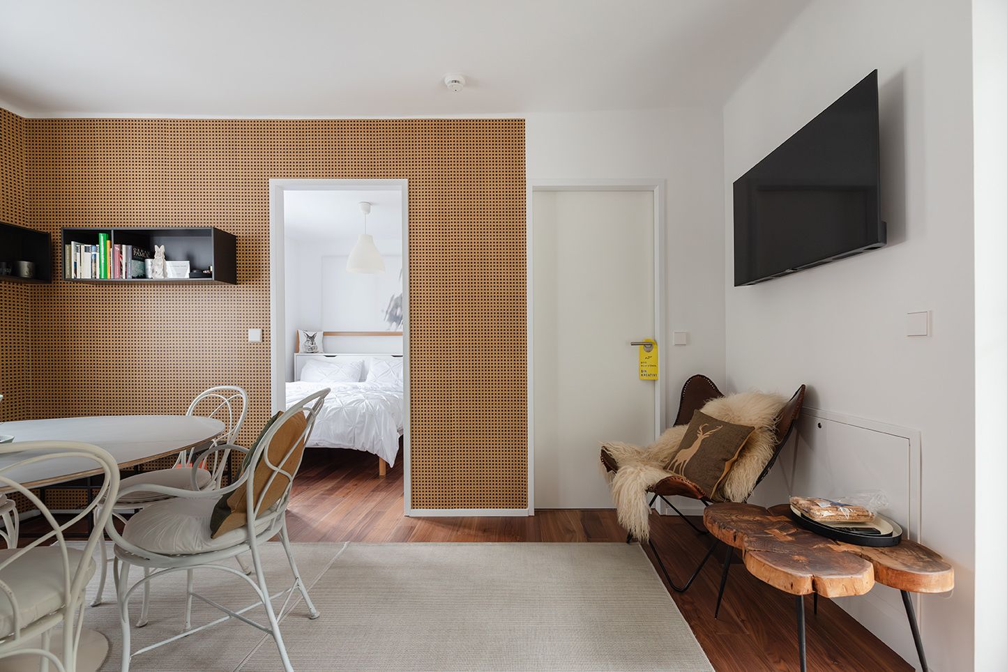 Modern eingerichtetes Zimmer mit Holzwand, Esstisch, Stühlen und einem Schlafzimmer im Hintergrund.
