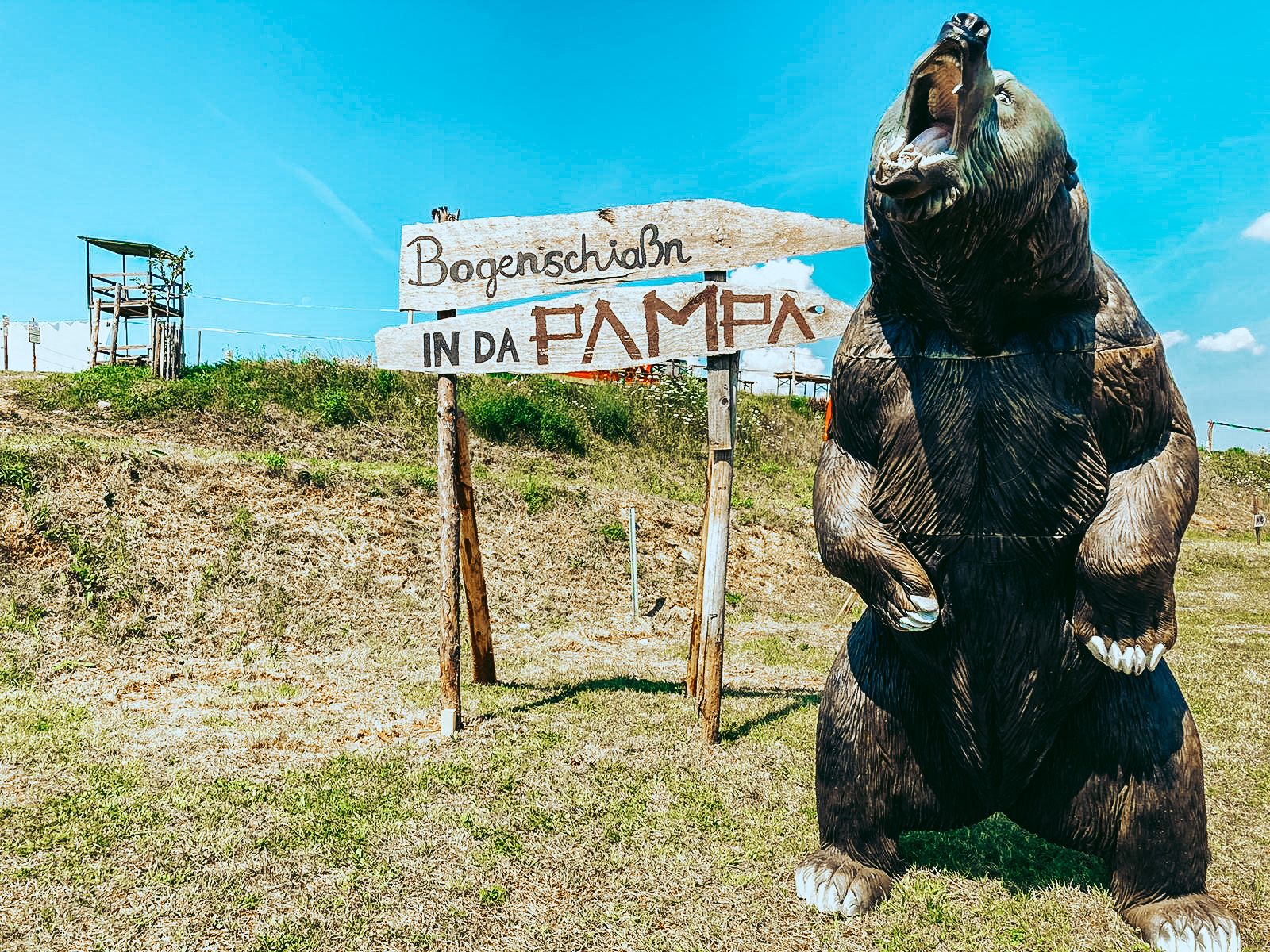 Ein großes Bärenmodell neben einem Schild mit der Aufschrift 'Bogenschiaßn in da Pampa' auf einer Wiese.