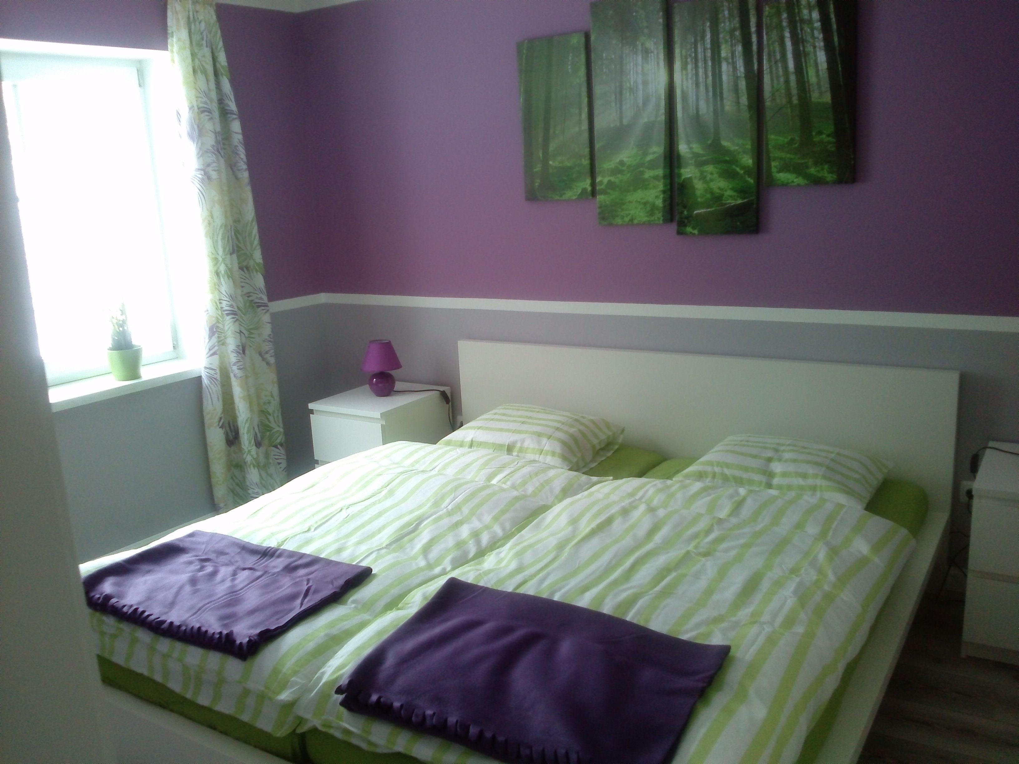 Schlafzimmer mit lila und grüner Dekoration, Doppelbett, Nachttisch und Fenster.