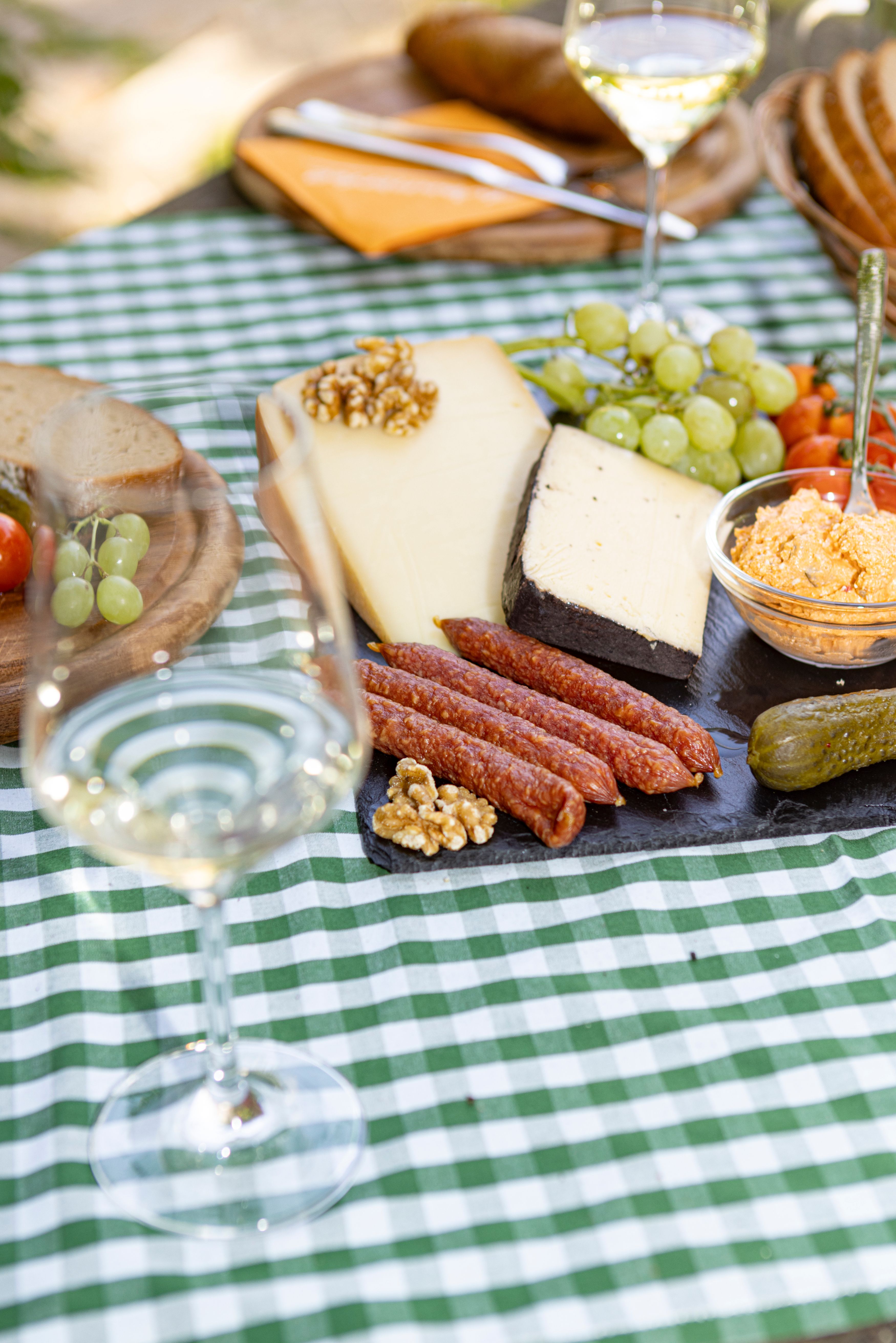Picknicktisch mit Käse, Wurst, Brot, Weintrauben und einem Glas Weißwein auf kariertem Tischtuch.