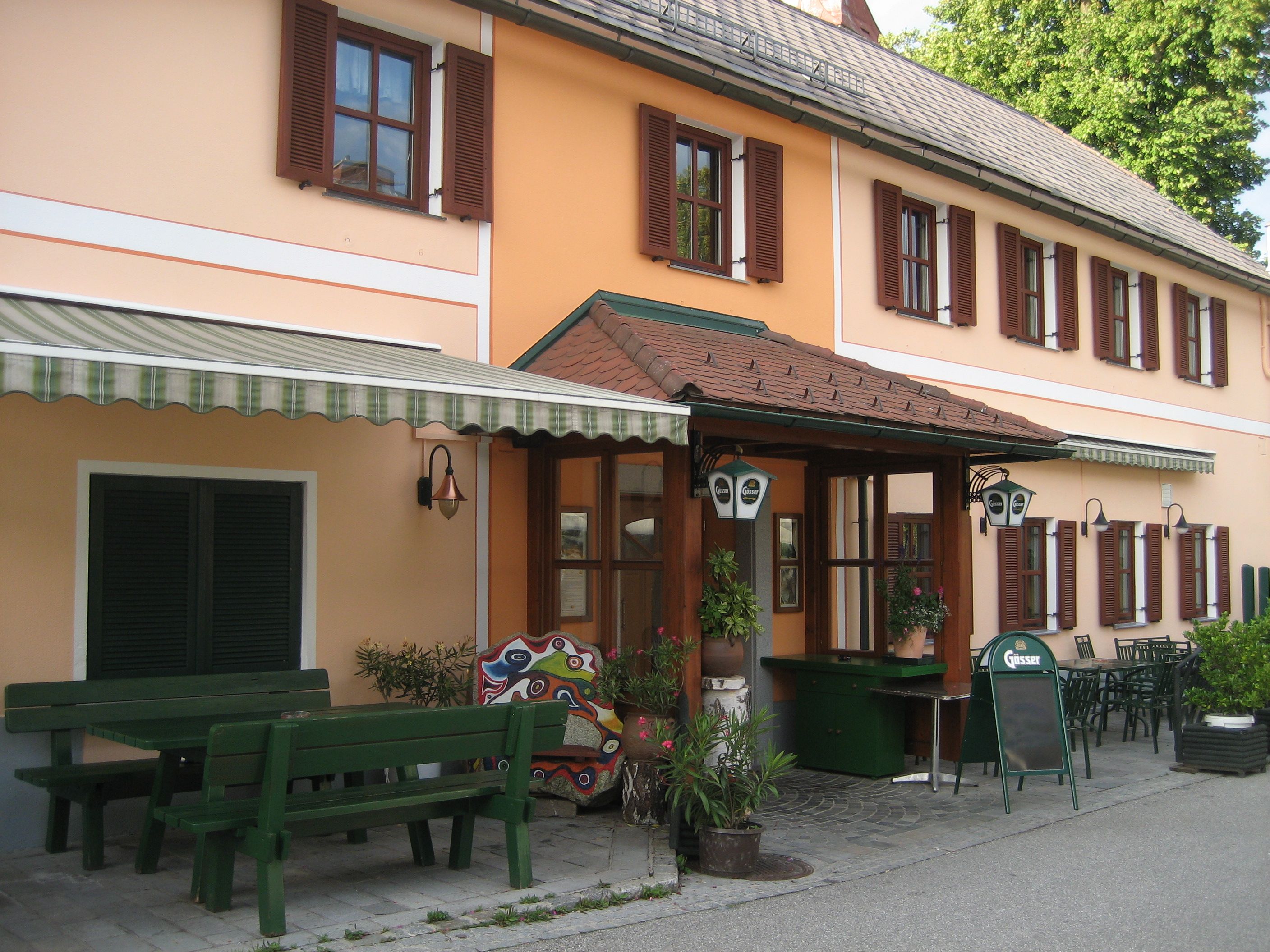 Eingang eines traditionellen Gasthofs mit orangefarbener Fassade und grünen Holzmöbeln im Freien.