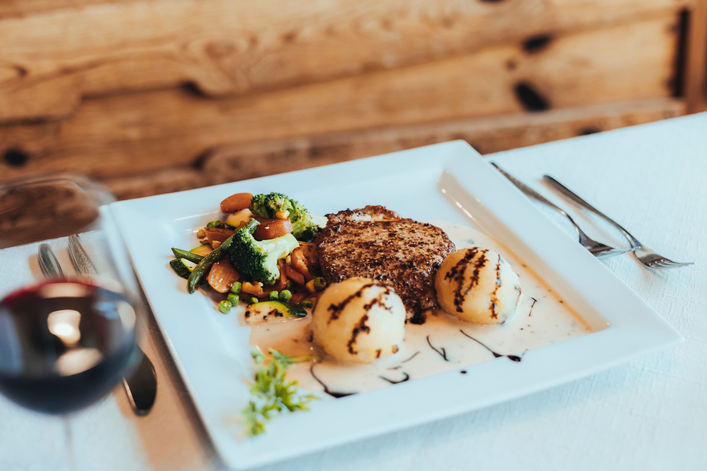 Ein Teller mit einem Kotelett, Gemüse und Knödel, garniert mit Soße und Balsamico, auf einem weißen Tisch.