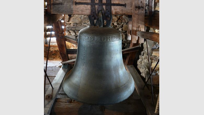 Große Glocke in einem hölzernen Glockenstuhl, umgeben von Steinmauern.